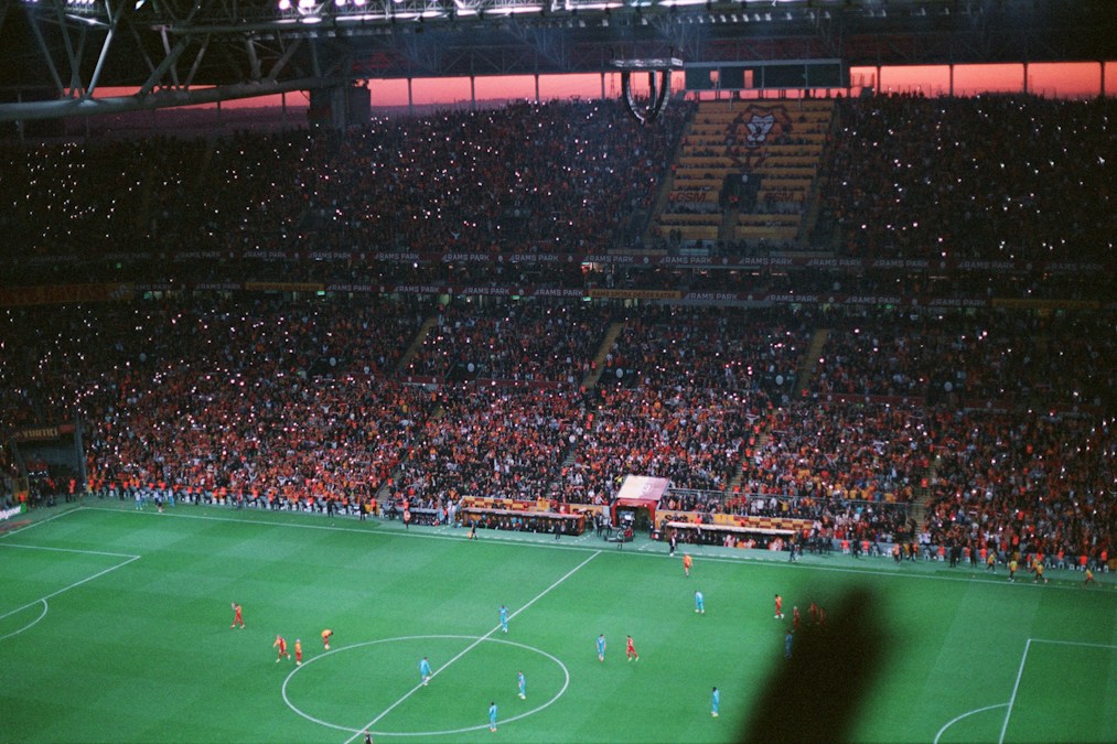 Un stade de football plein au crépuscule, tribunes rouges et orange piquetées des lumières des téléphones, joueurs sur la pelouse
