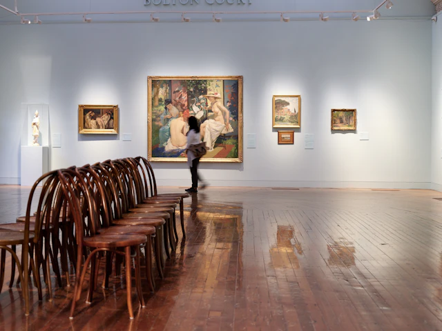 A gallery room with wooden benches and large paintings on pale walls