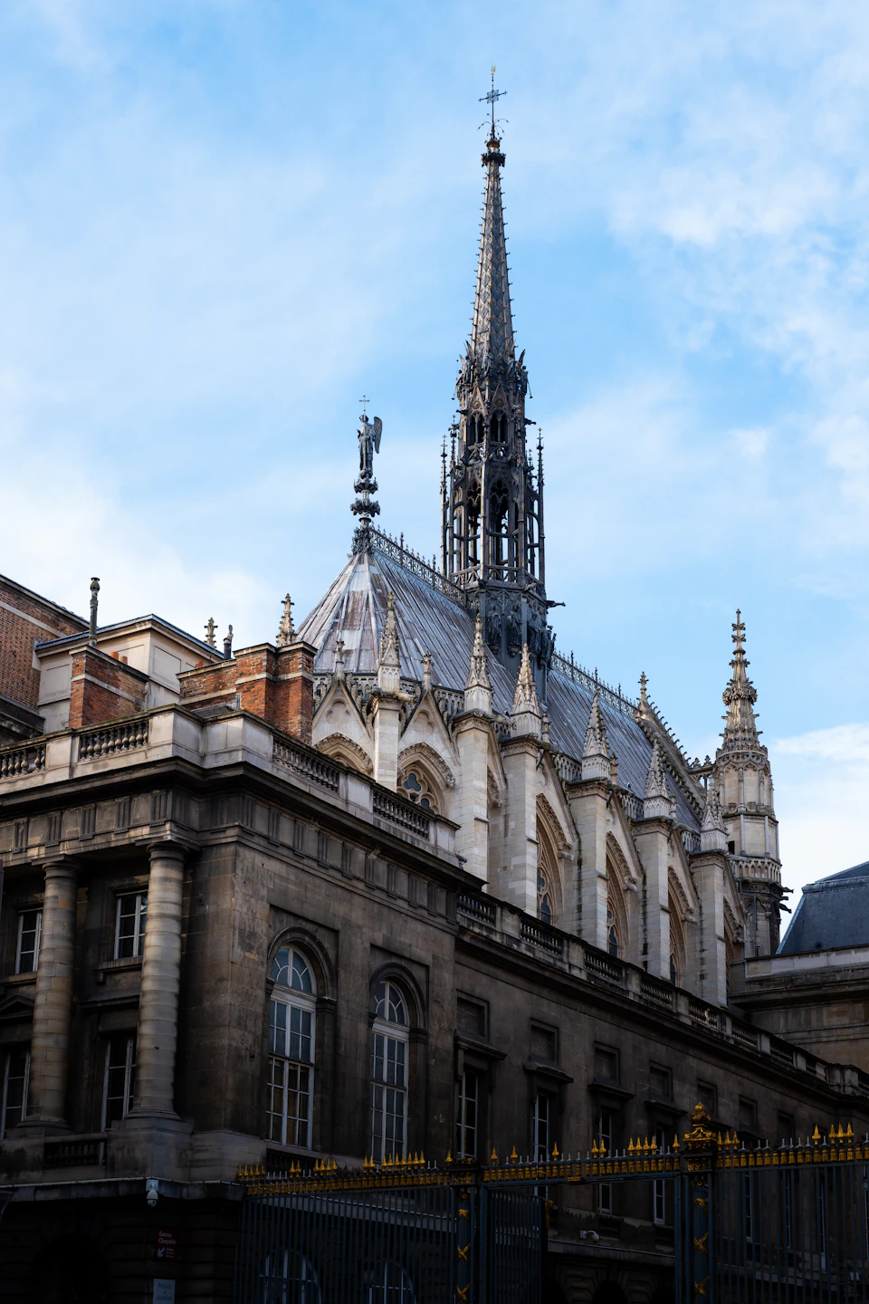 The image depicts a large, ornate stone building with multiple spires and towers reaching towards the sky. The building is situated on a street corner, surrounded by other structures that create an urban landscape. A statue can be seen atop one of the buildings, adding to its architectural charm. The perspective of the photo is taken from below, looking up at the imposing height of the structure.