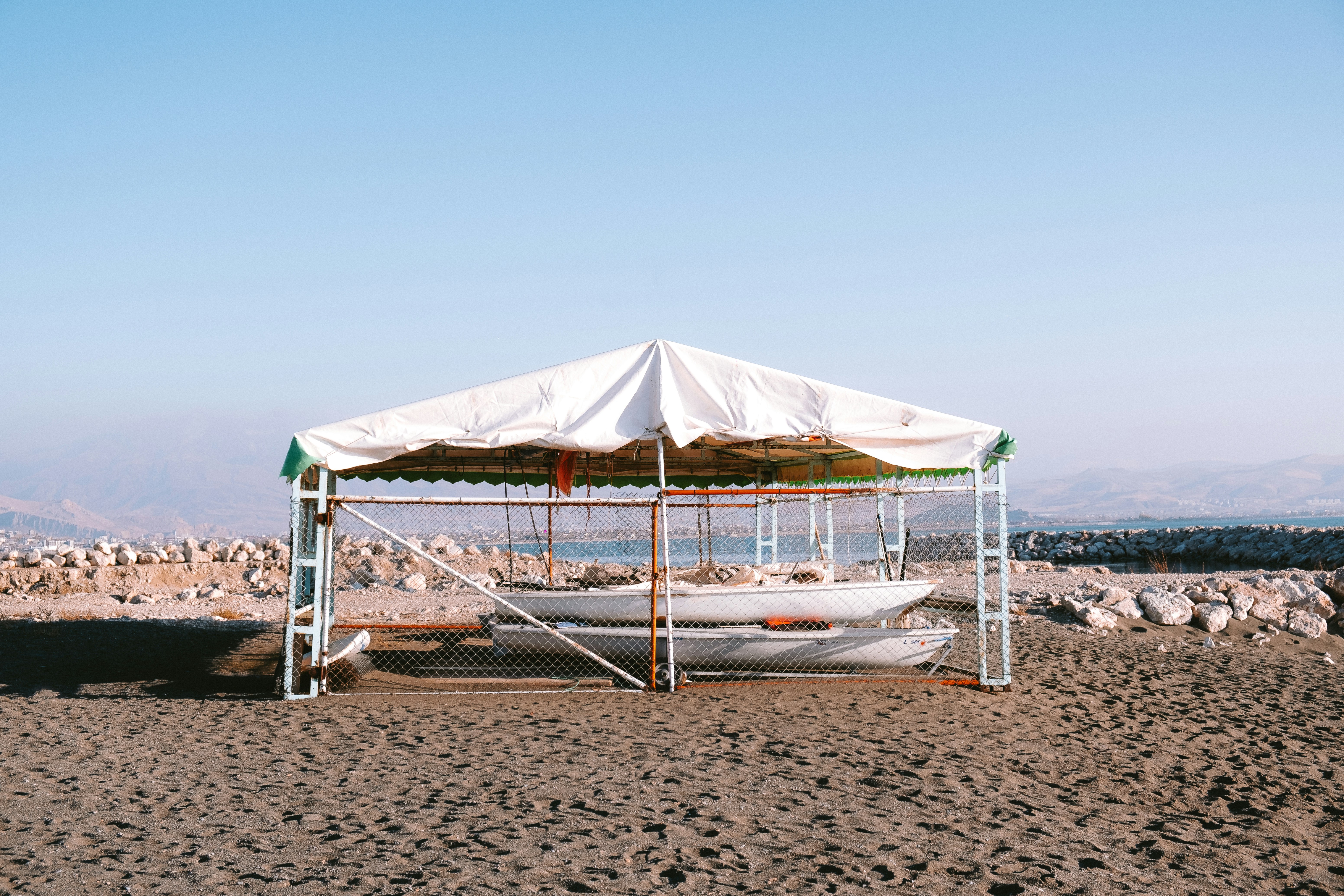 White canopy on a metal frame on a sandy beach, small boats stored underneath and rocky hills behind