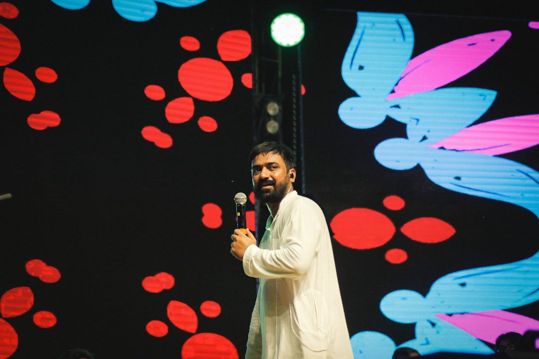 Actor-politician Vijay addressing a large crowd at a political rally, wearing a simple white shirt and black trousers, with TVK party flags in the background.