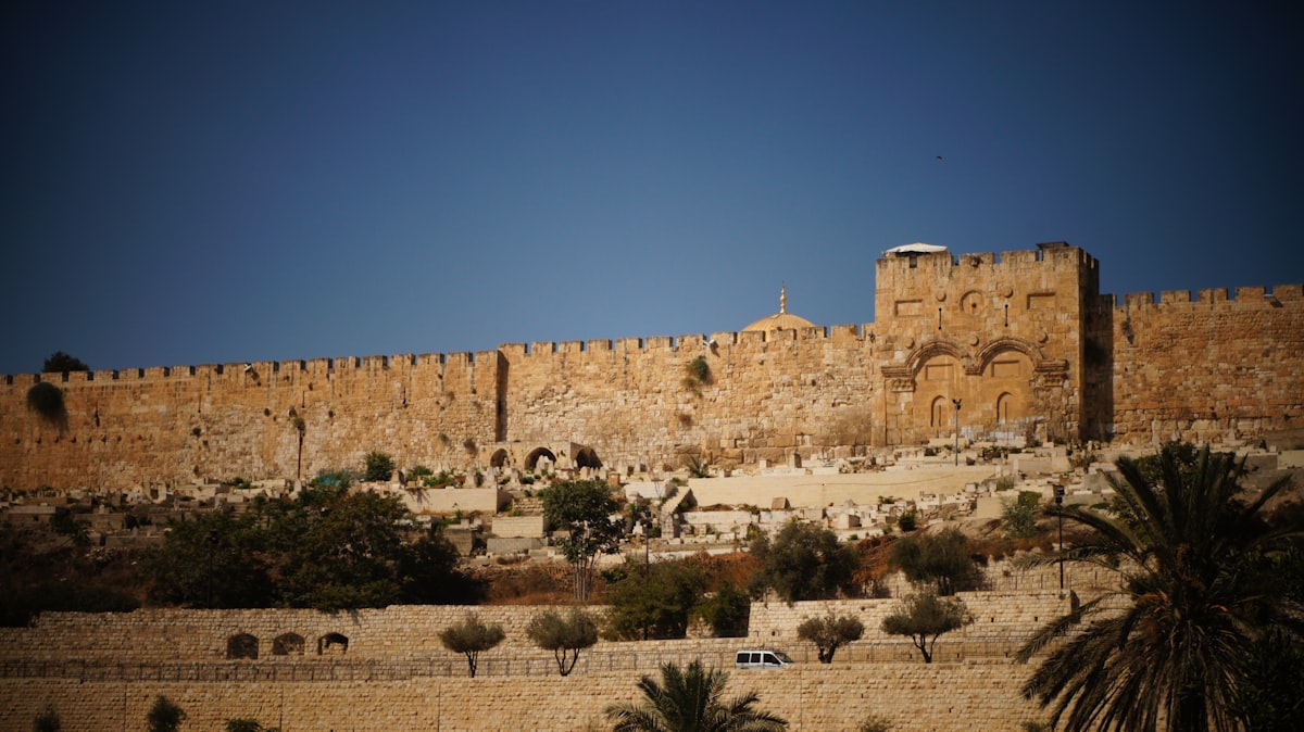 The stone walls of the Old City of Jerusalem in bright daylight, with an arched gate and a watchtower above them, and the roofs and towers of the city behind under a blue sky