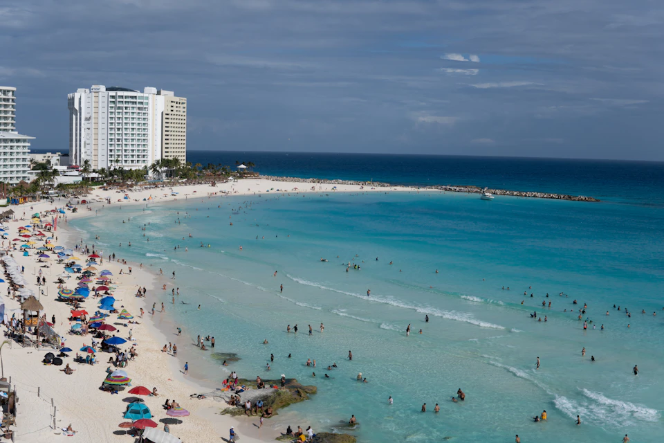 The image depicts a crowded beach scene with numerous people enjoying their time on the sandy shoreline, which is dotted with umbrellas and lounge chairs. The azure ocean water gently laps against the shore, creating small waves that add to the lively atmosphere of the beach. In the distance, several tall buildings can be seen, suggesting that this beach is located near a city or urban area.