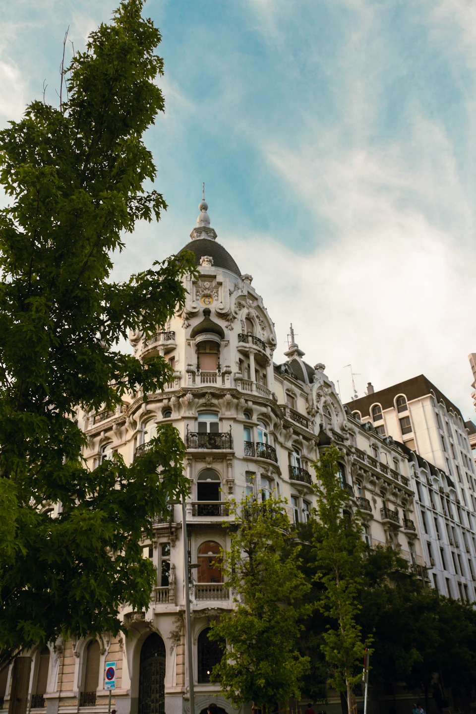 The image depicts a large, ornate white building with a dome and multiple levels, situated on a street corner. The building is surrounded by lush green trees, creating a contrast between the architectural structure and nature's elements. In front of the building, there are two smaller buildings visible in the background.