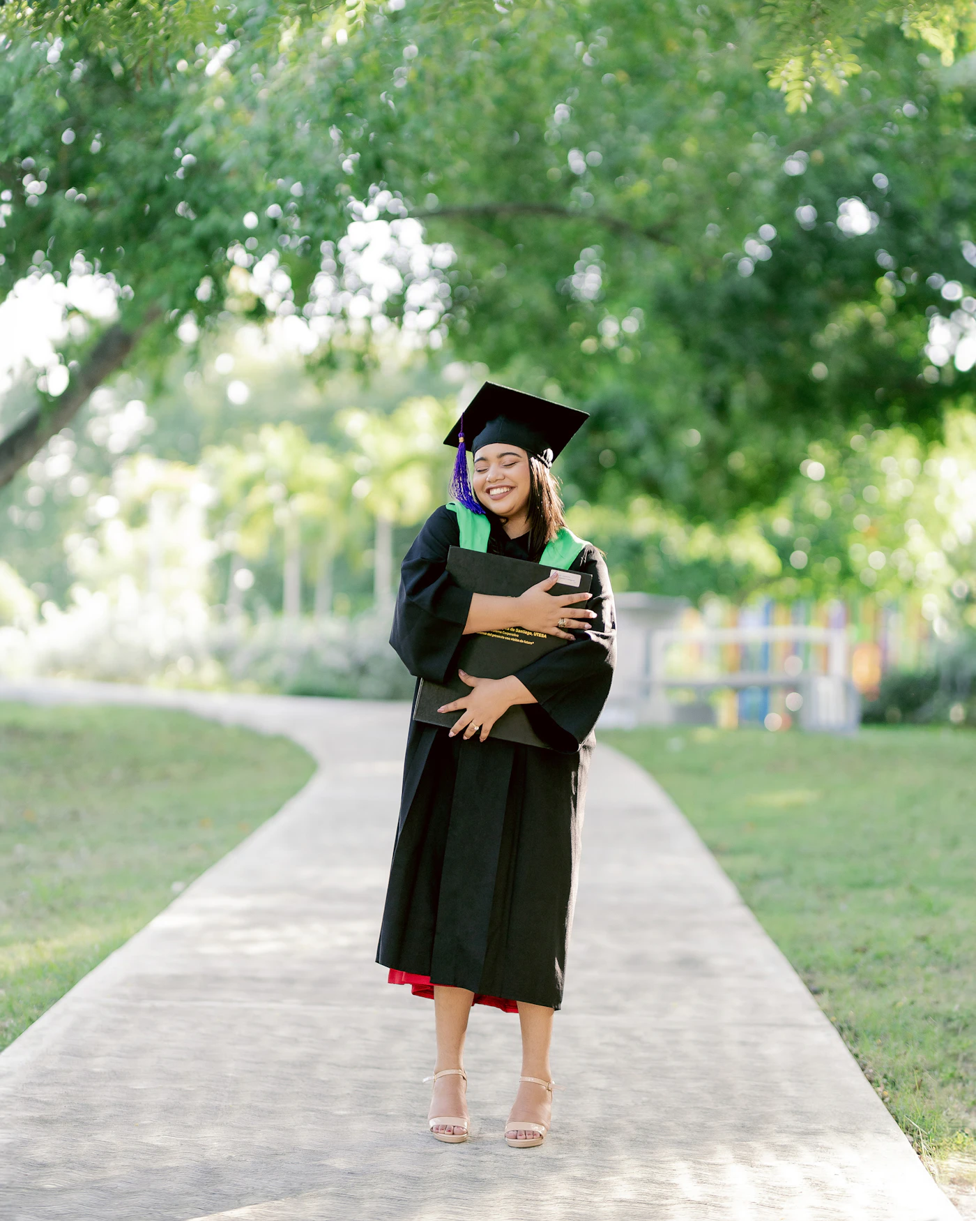 A young graduate in a cap and gown standing on a campus sidewalk on graduation day
