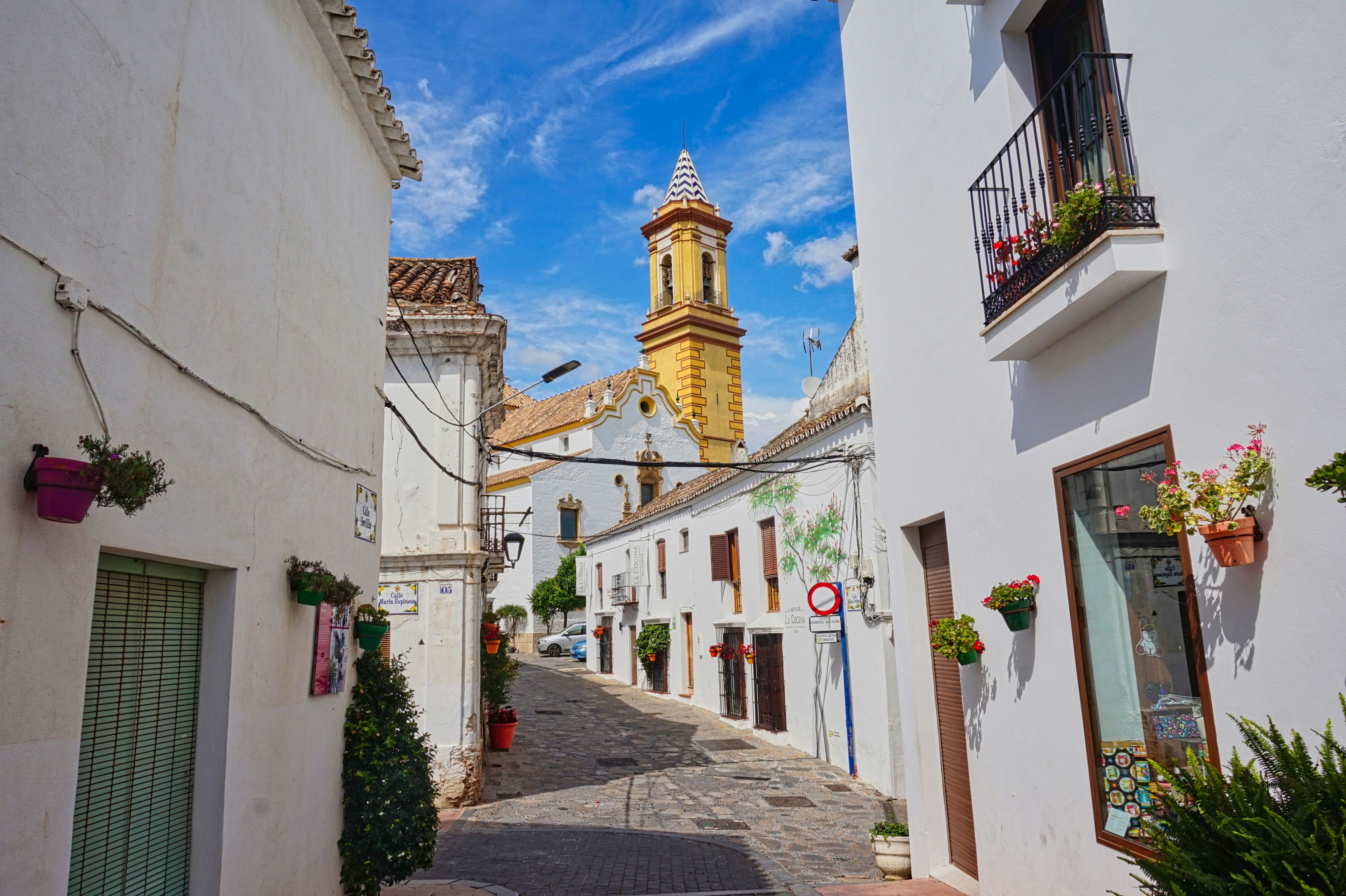 A narrow street with a clock tower in the background, An alley in Estepona with view of a church in the background