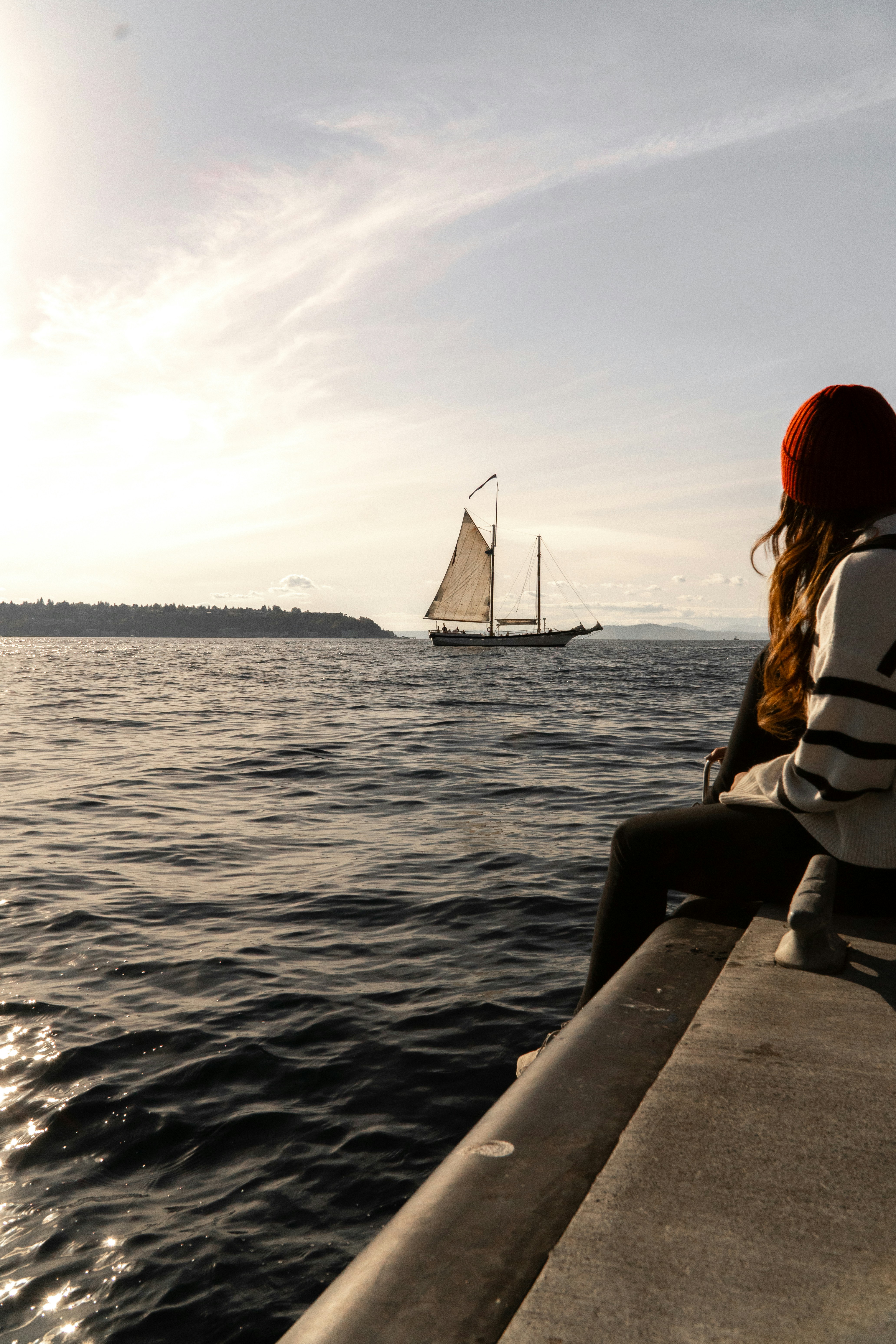 A person sitting on the edge of a pier looking at a sailboat