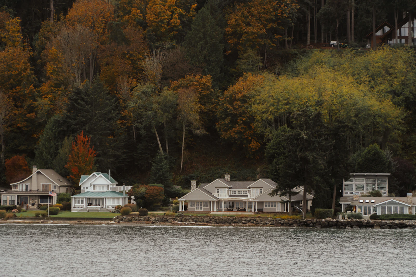 Row of waterfront houses at the shoreline below a steep hillside of autumn-colored evergreen forest
