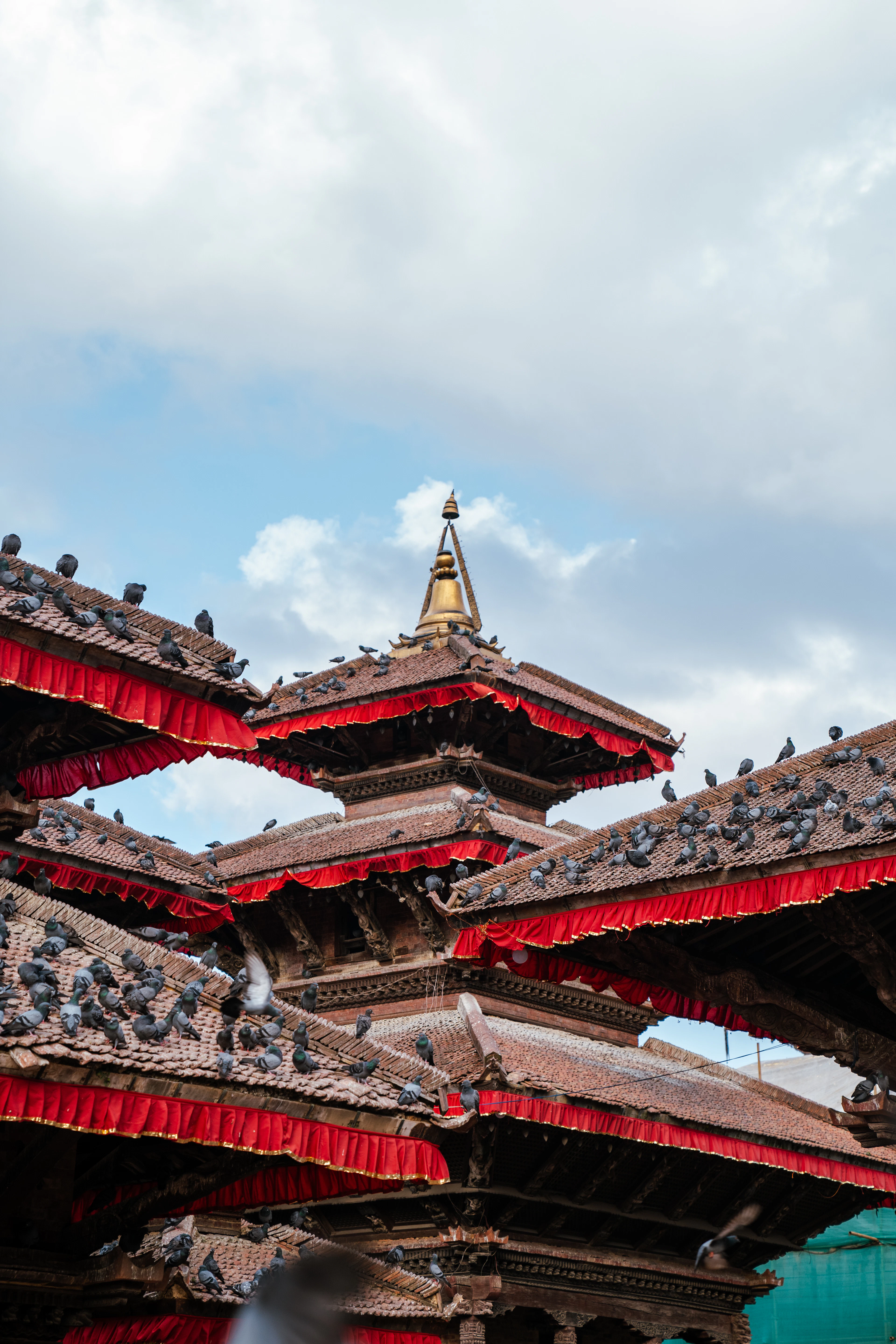 Tiered pagoda roofs at Kathmandu Durbar Square, their eaves hung with scarlet cloth valances and crowded with roosting pigeons, a gilded finial rising into a pale cloudy sky