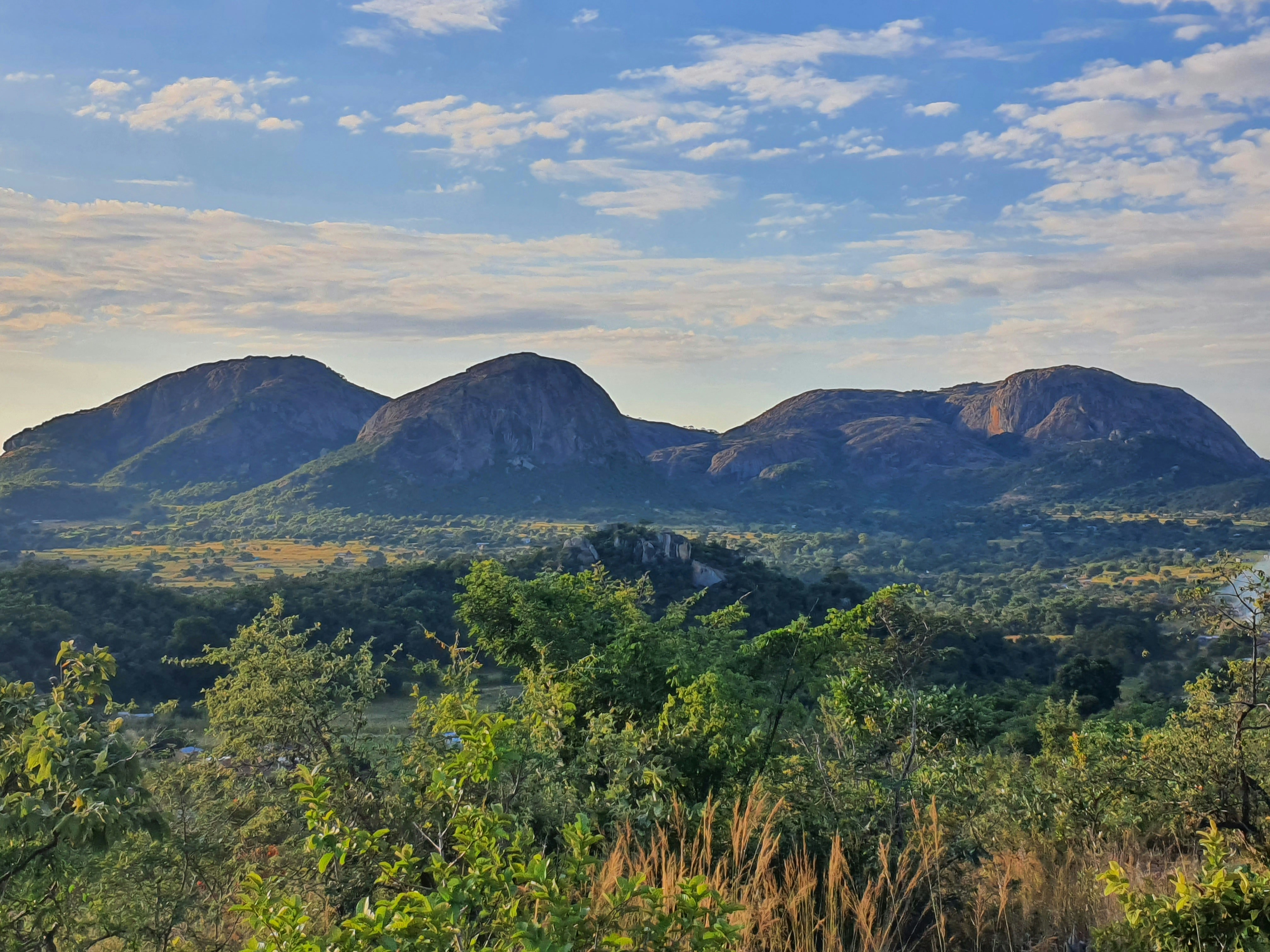 A scenic view of a mountain range in the distance, Ngomakurira, situated atop the soils of Domboshava in Zimbabwe, is a majestic granite mountain steeped in cultural, historical, and spiritual significance. The mountain is adorned with smooth, bare rock faces and dotted with patches of hardy vegetation, including indigenous shrubs and trees that cling tenaciously to cracks and crevices. Its rugged beauty is accentuated by its reddish-gray hues, particularly striking during sunrise and sunset.