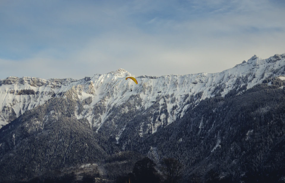 The image depicts a snow-capped mountain range under a clear blue sky with fluffy white clouds. A yellow paraglider is soaring high above the mountains, adding a vibrant contrast to the serene landscape. The paraglider's position in the top right corner of the frame suggests it has reached its peak and is now descending towards the lower part of the image.

The mountain range itself is a majestic sight with snow-covered peaks that stretch across the entire background, creating a sense of depth and scale. The clear blue sky above adds to the overall beauty of the scene.