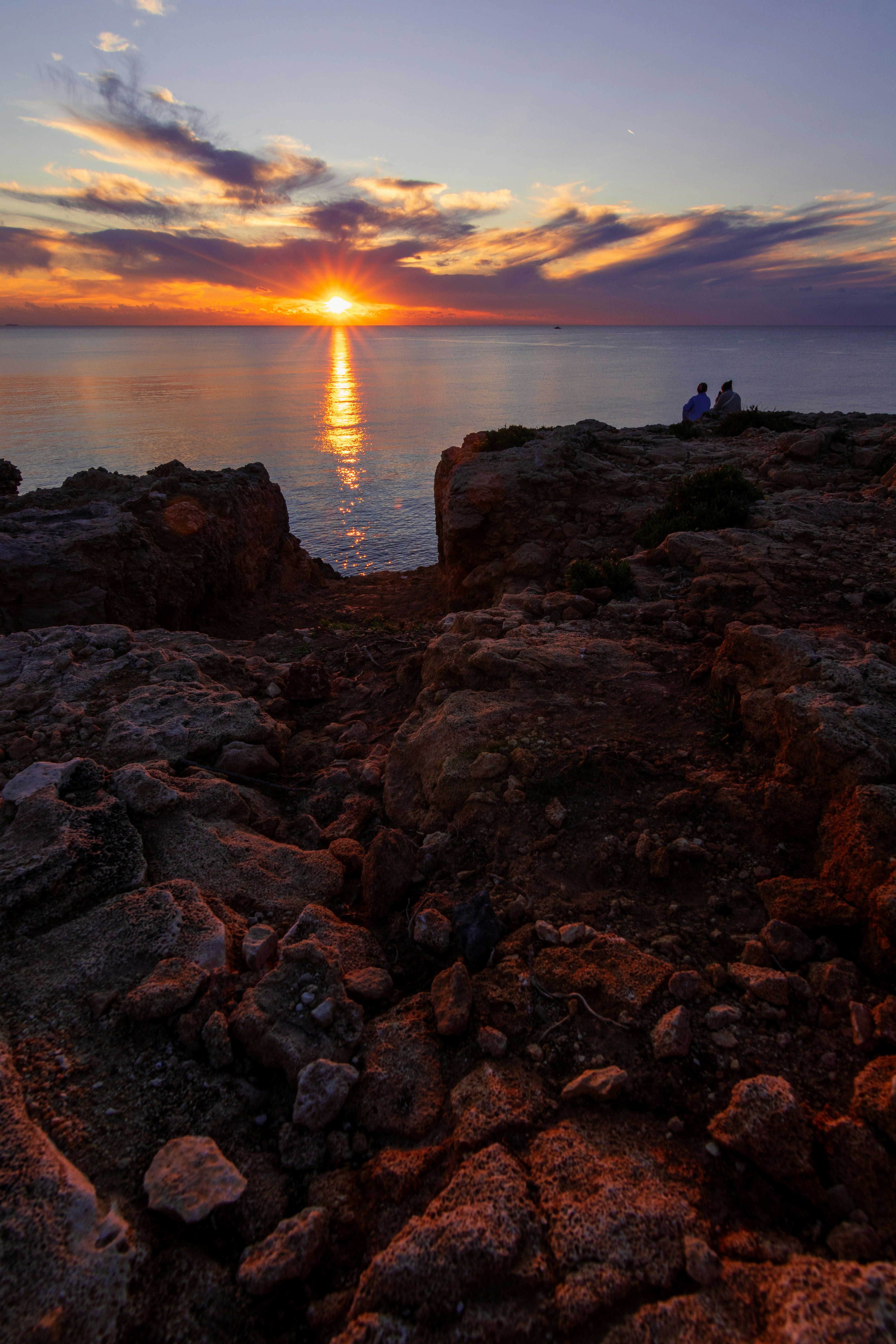 A couple of people are sitting on a rock by the water