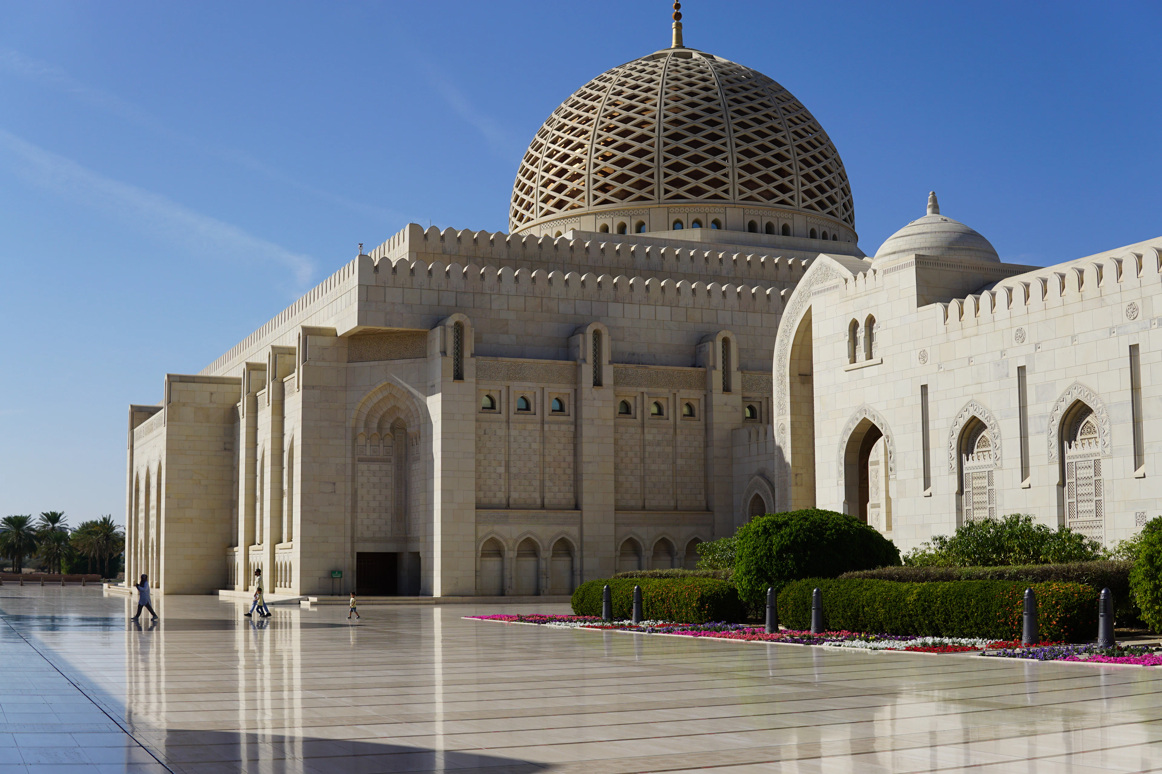The Sultan Qaboos Grand Mosque's ribbed dome and minaret rise above manicured gardens and red flowerbeds.