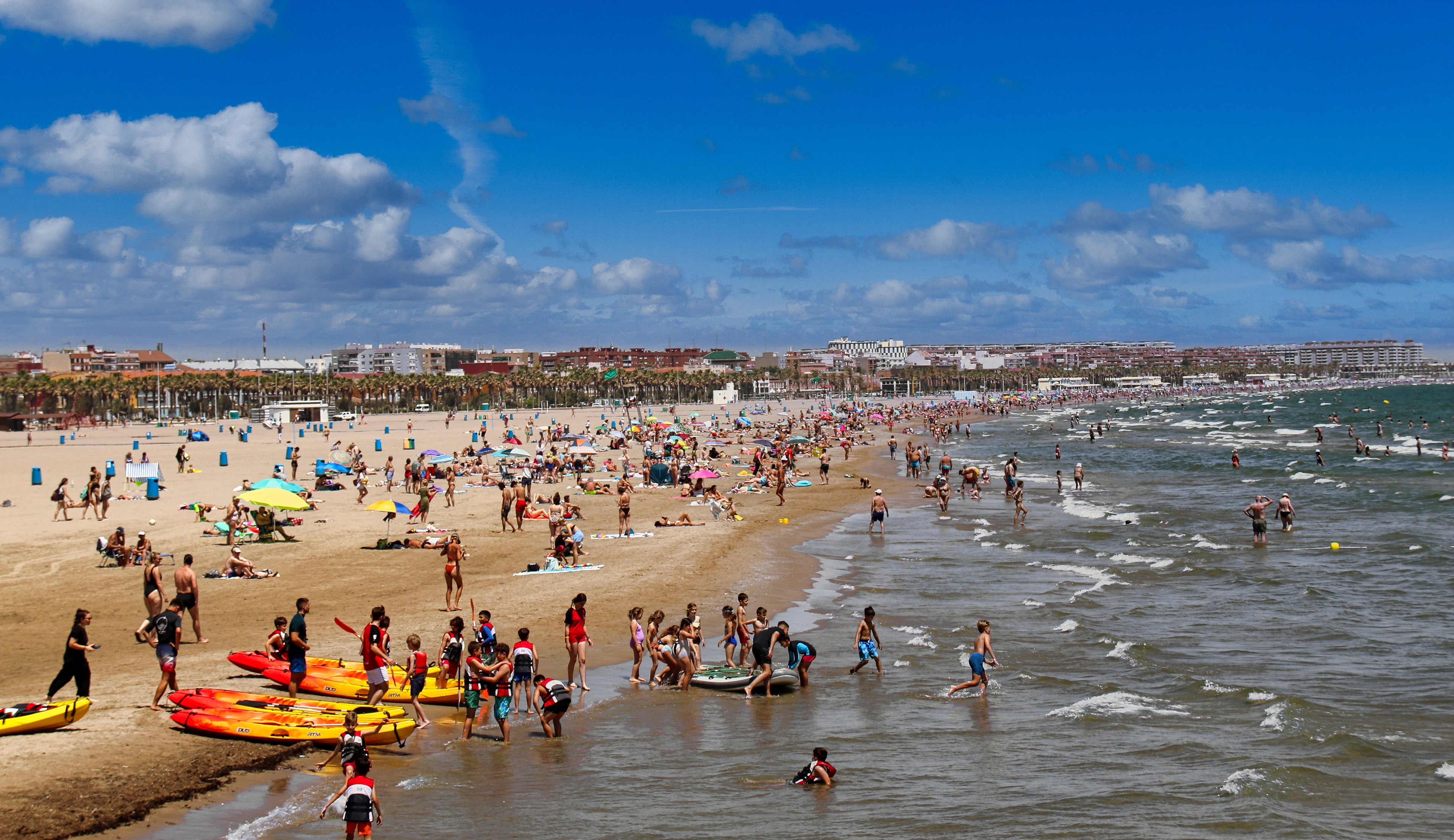 Sunbathers, kayaks and swimmers crowd a lively Valencia beach under a clear summer sky.