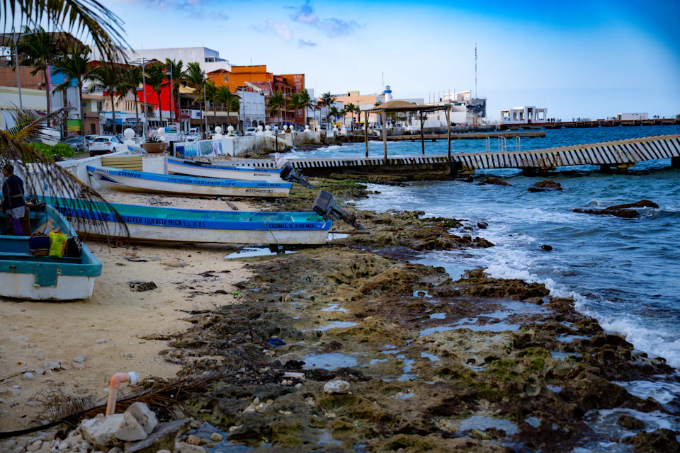The image depicts a serene beach scene with several boats moored along the shoreline, including blue and white boats and a green boat. The boats are neatly arranged on the sandy beach, facing towards the water. In the background, there is a pier extending into the ocean, which adds to the picturesque setting. The sky above is clear and blue, suggesting a pleasant day at the beach.