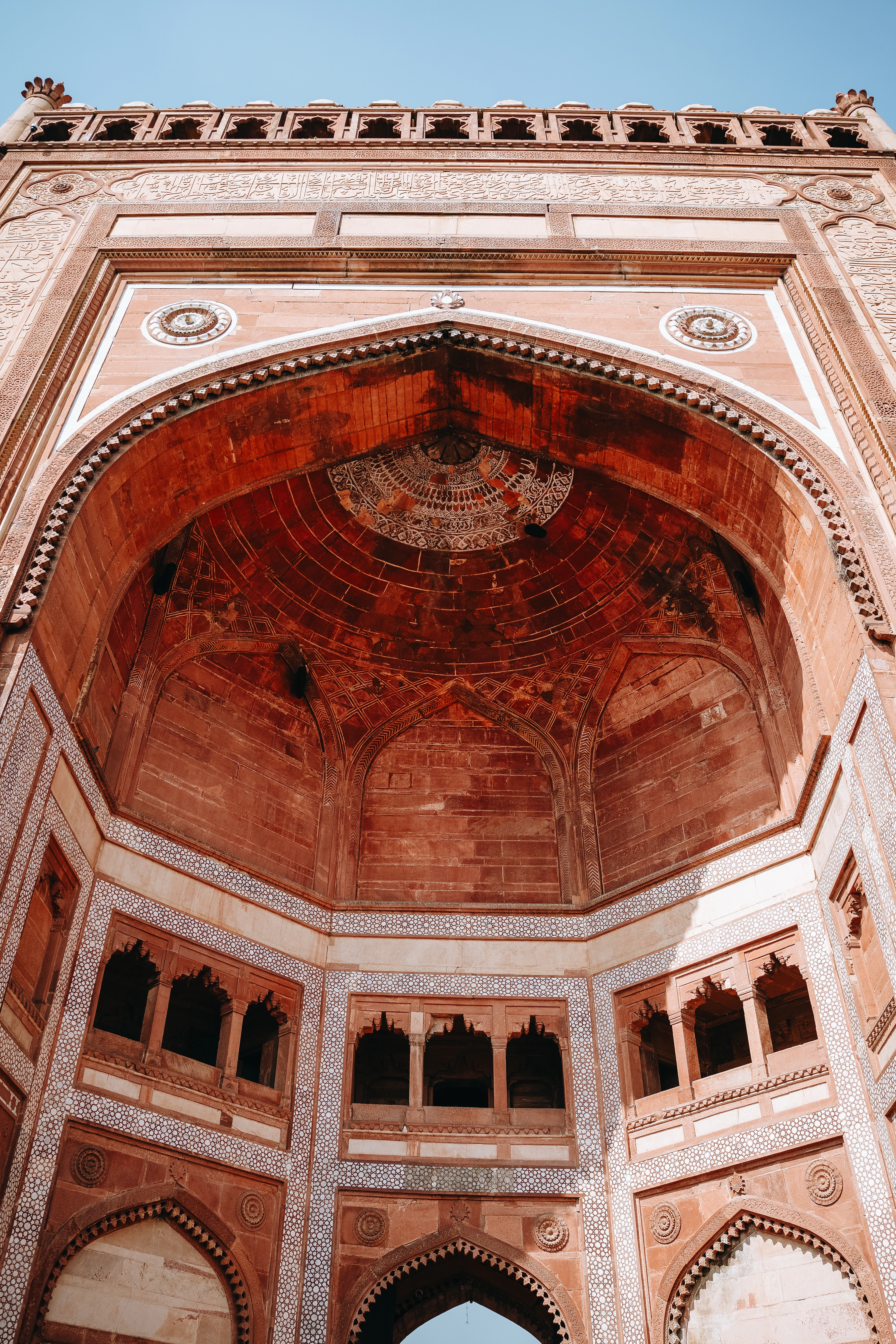 The Buland Darwaza at Fatehpur Sikri seen from directly below: a colossal red sandstone gateway banded with white marble inlay, its half-dome arch and rows of carved niches rising into a clear blue sky.