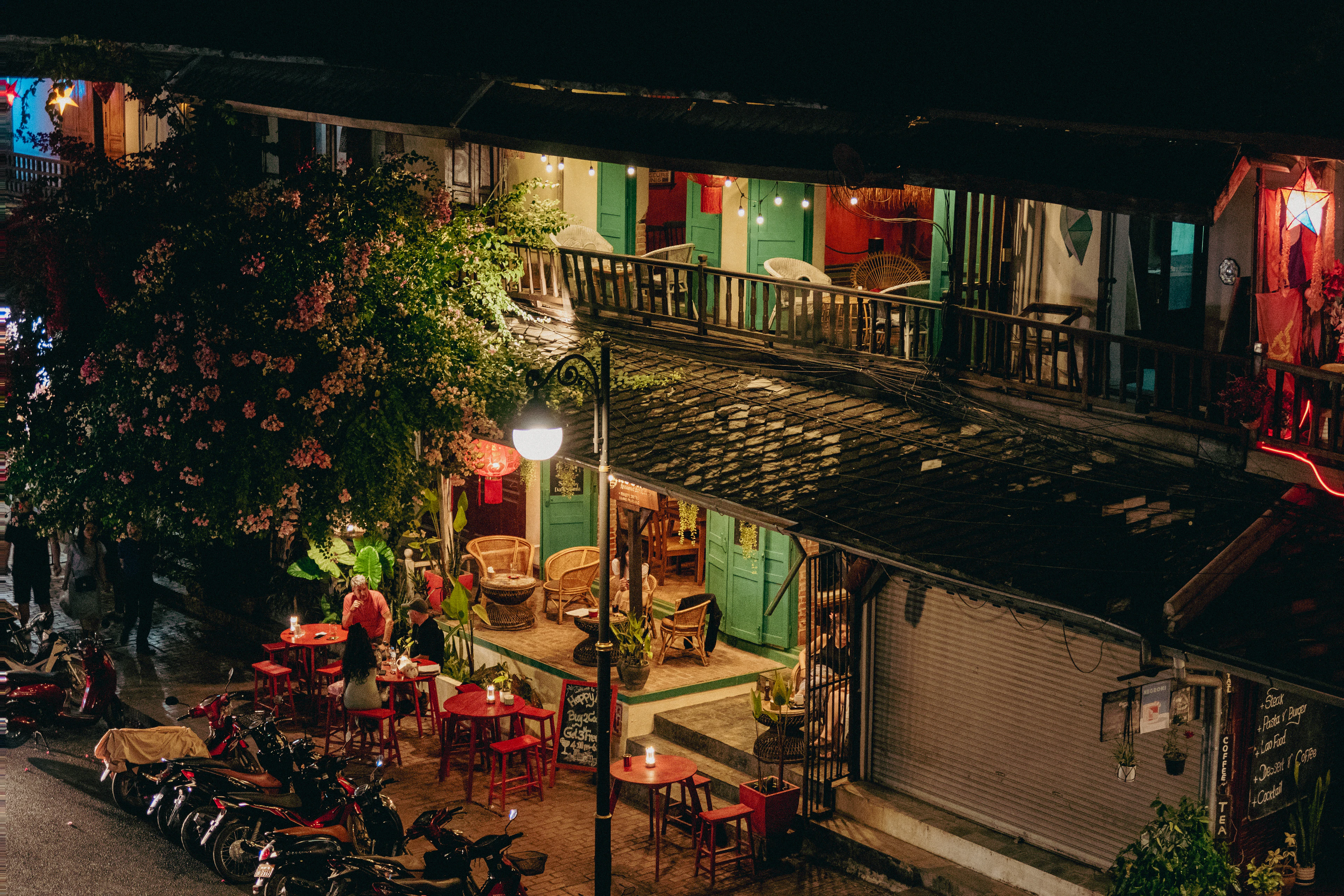 A near-empty street in Luang Prabang at night, warm light spilling from a green-shuttered colonial shophouse onto rattan chairs and small red tables under a flowering tree