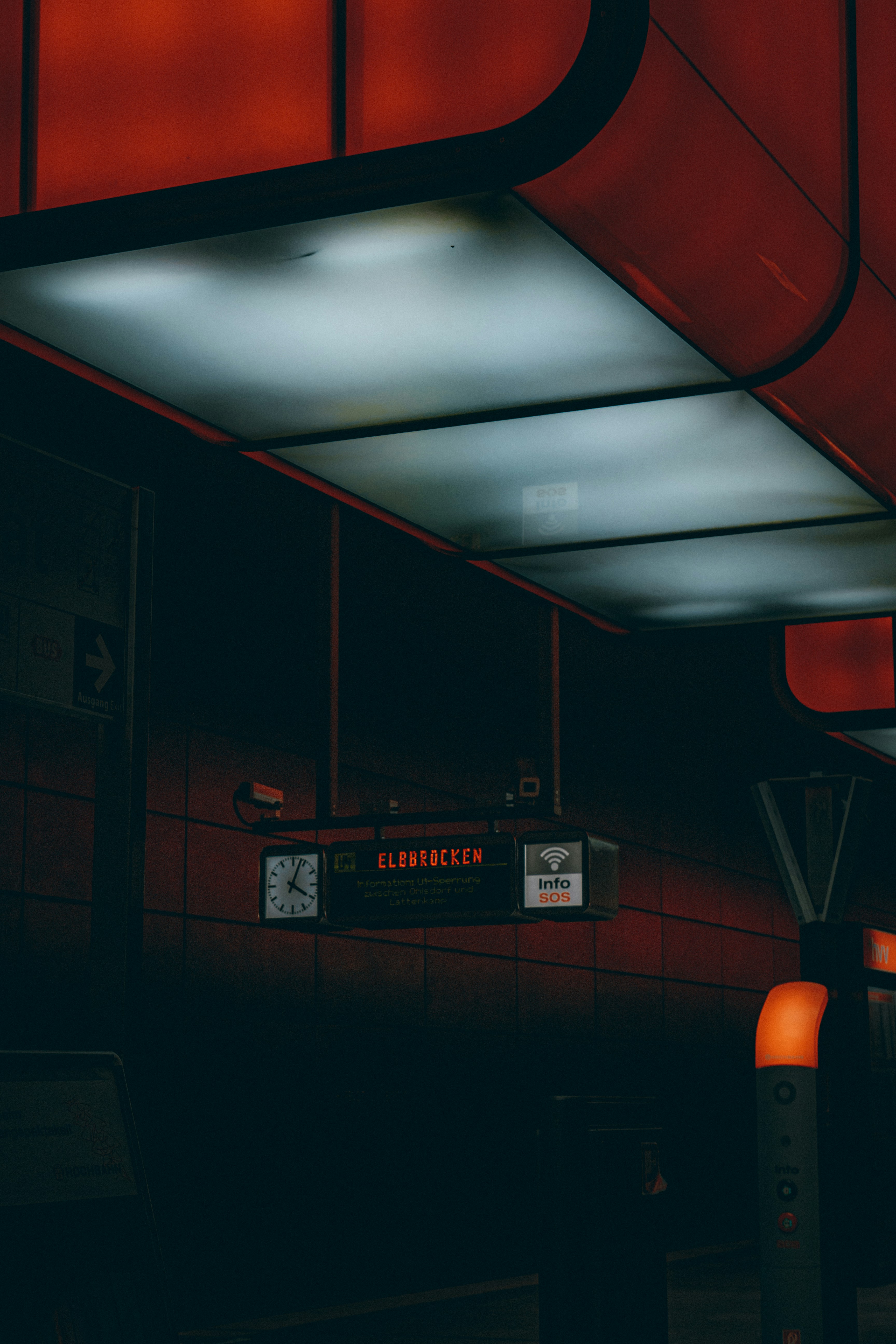 Dimly lit street-level photograph of a bus stop under a red canopy, featuring a glowing digital sign and a clock.
