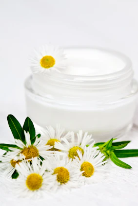 A jar of cream and some daisies on a white surface