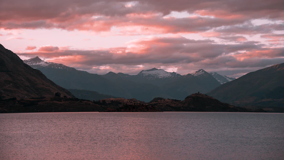 The image depicts a serene mountain landscape at sunset, with a large body of water reflecting the warm hues of the setting sun. The sky is painted in shades of pink and orange, creating a stunning backdrop for the scene. In the distance, several snow-capped mountains rise majestically against this colorful sky. The perspective of the image is from the shoreline, looking out towards the mountains, giving a sense of depth and vastness to the landscape.
