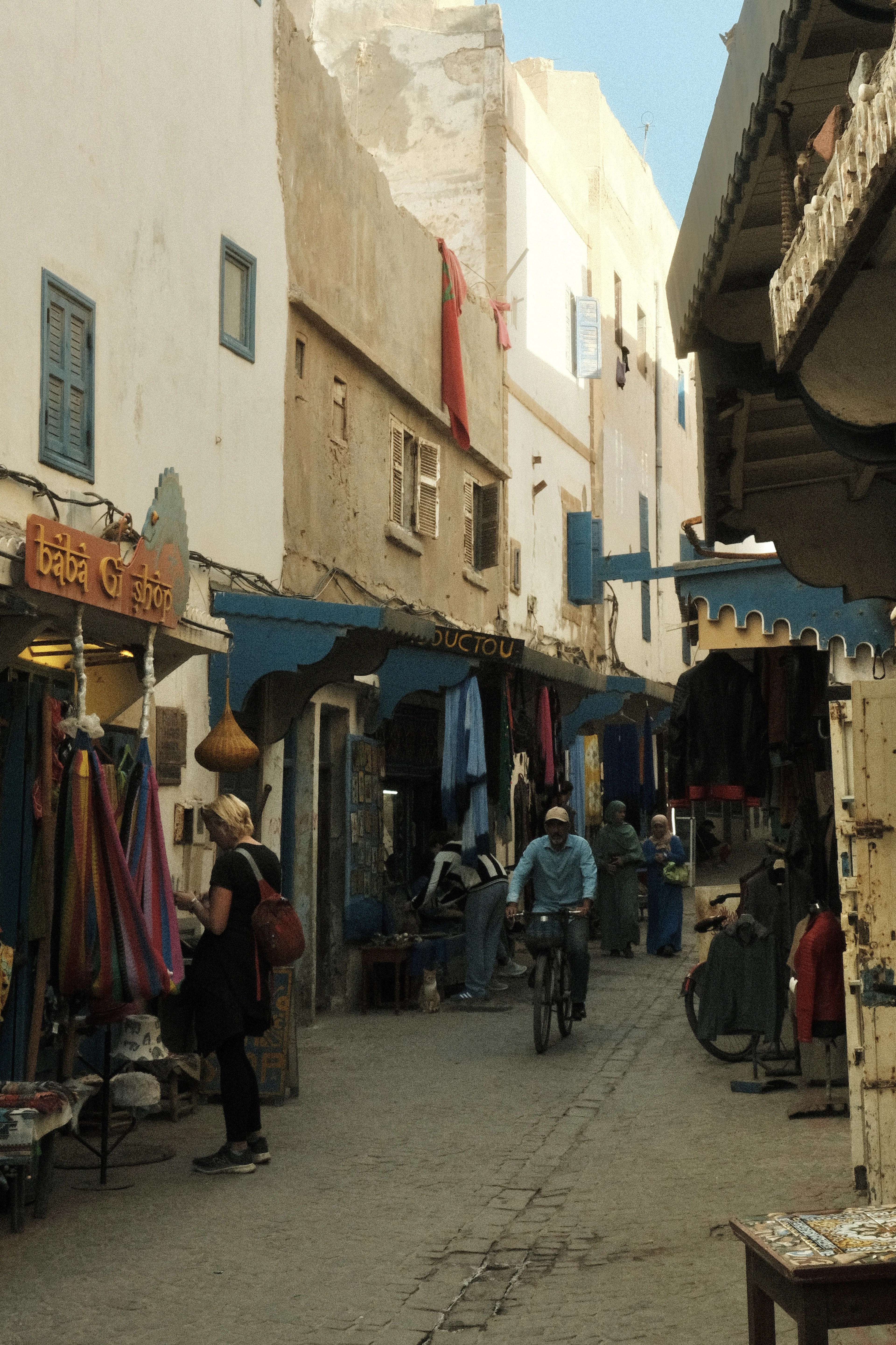 A cyclist rides through a narrow Essaouira medina alley lined with shops.