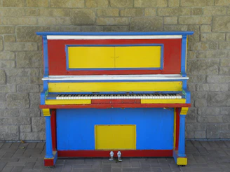 A red, yellow, and blue piano sitting in front of a brick wall