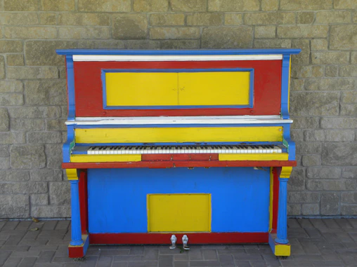 A red, yellow, and blue piano sitting in front of a brick wall
