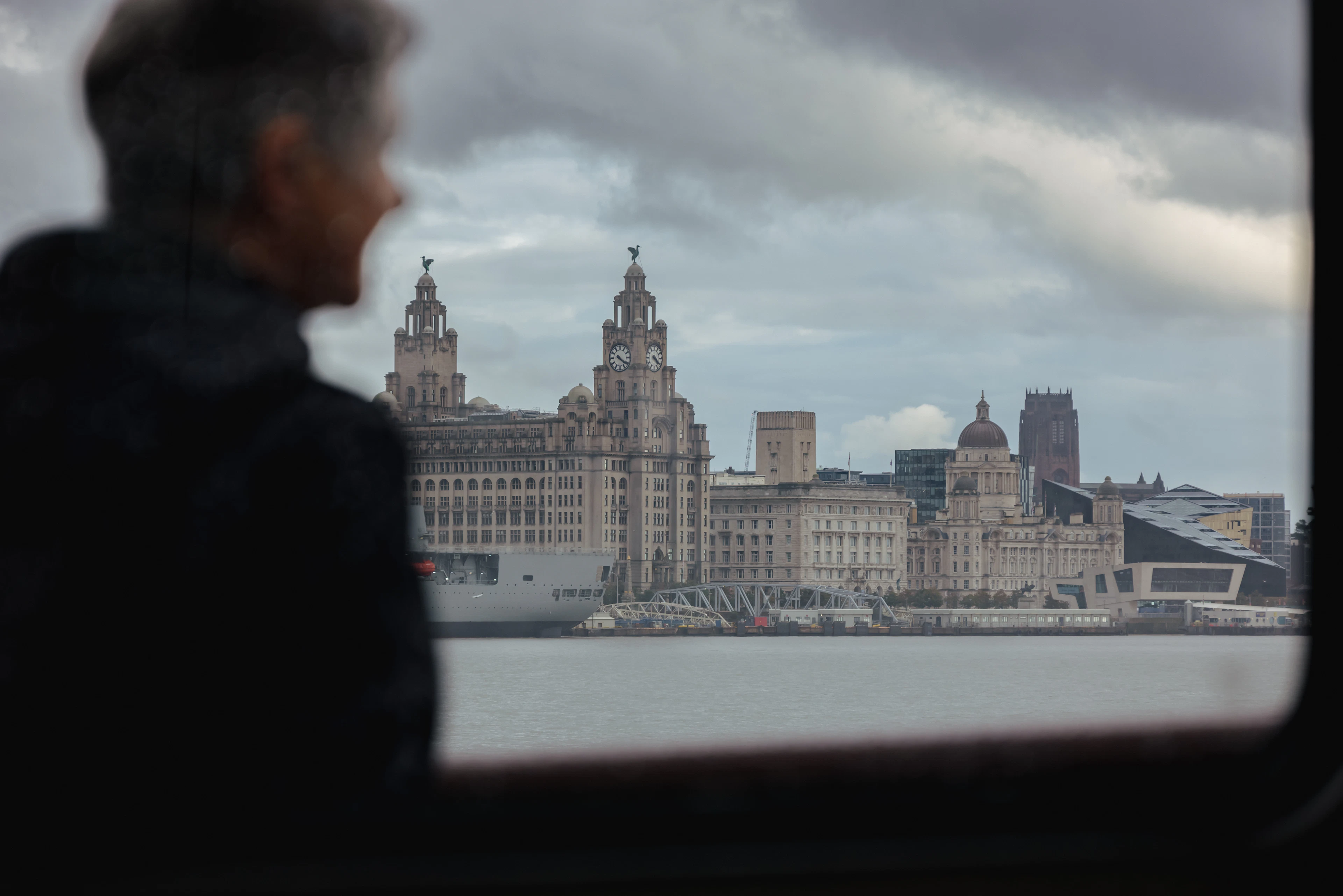 A ferry passenger's silhouette looking out at Liverpool's waterfront skyline across the Mersey under a moody sky.