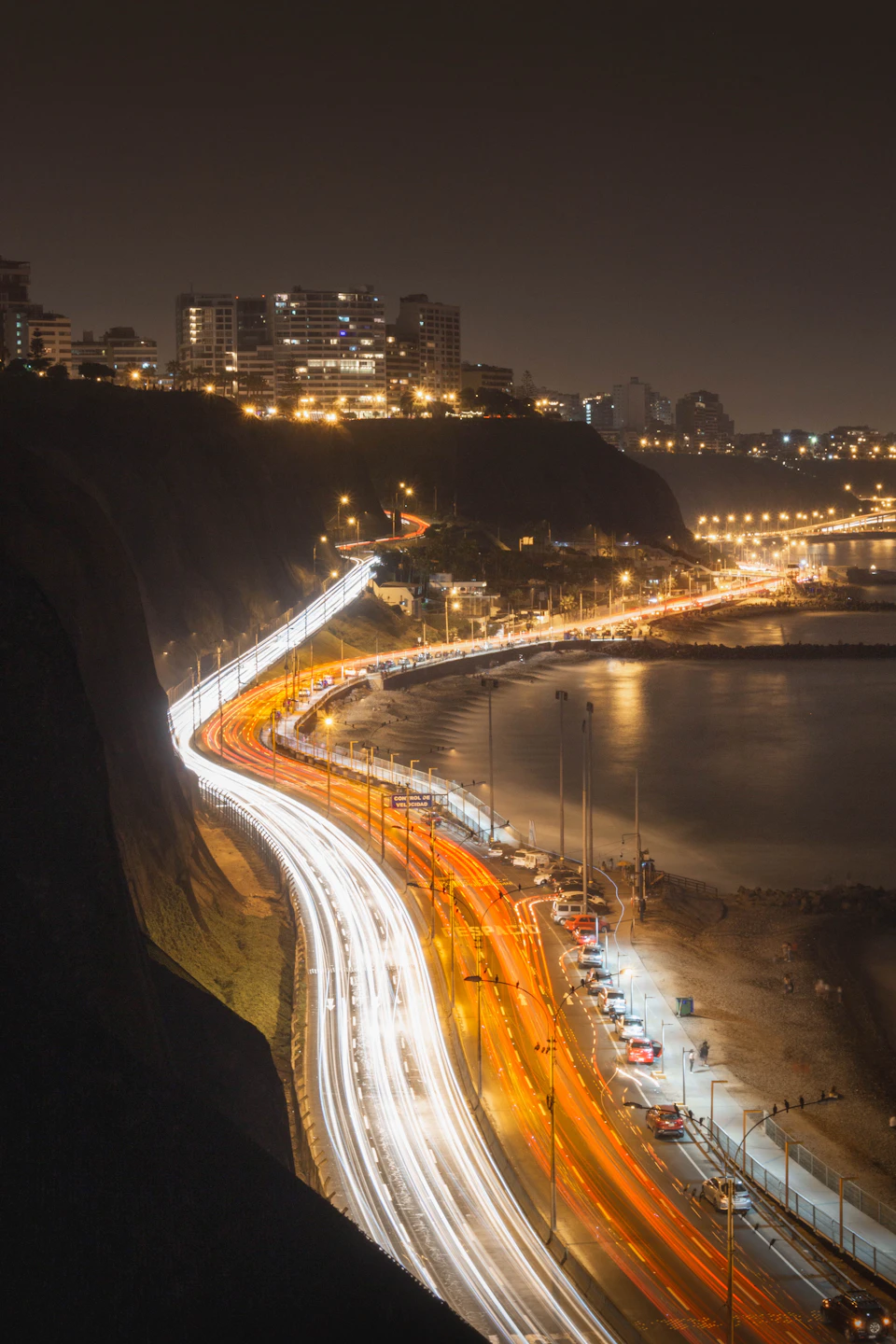 The image depicts a cityscape at night, with a winding road cutting through the scene. The road is illuminated by bright lights and features multiple lanes filled with cars moving in both directions. In the background, buildings rise against the dark sky, their windows twinkling like stars. A body of water can be seen on the right side of the image, adding to the overall ambiance of the city at night. The scene is captured from a high vantage point, providing a comprehensive view of the urban landscape and its various elements.
