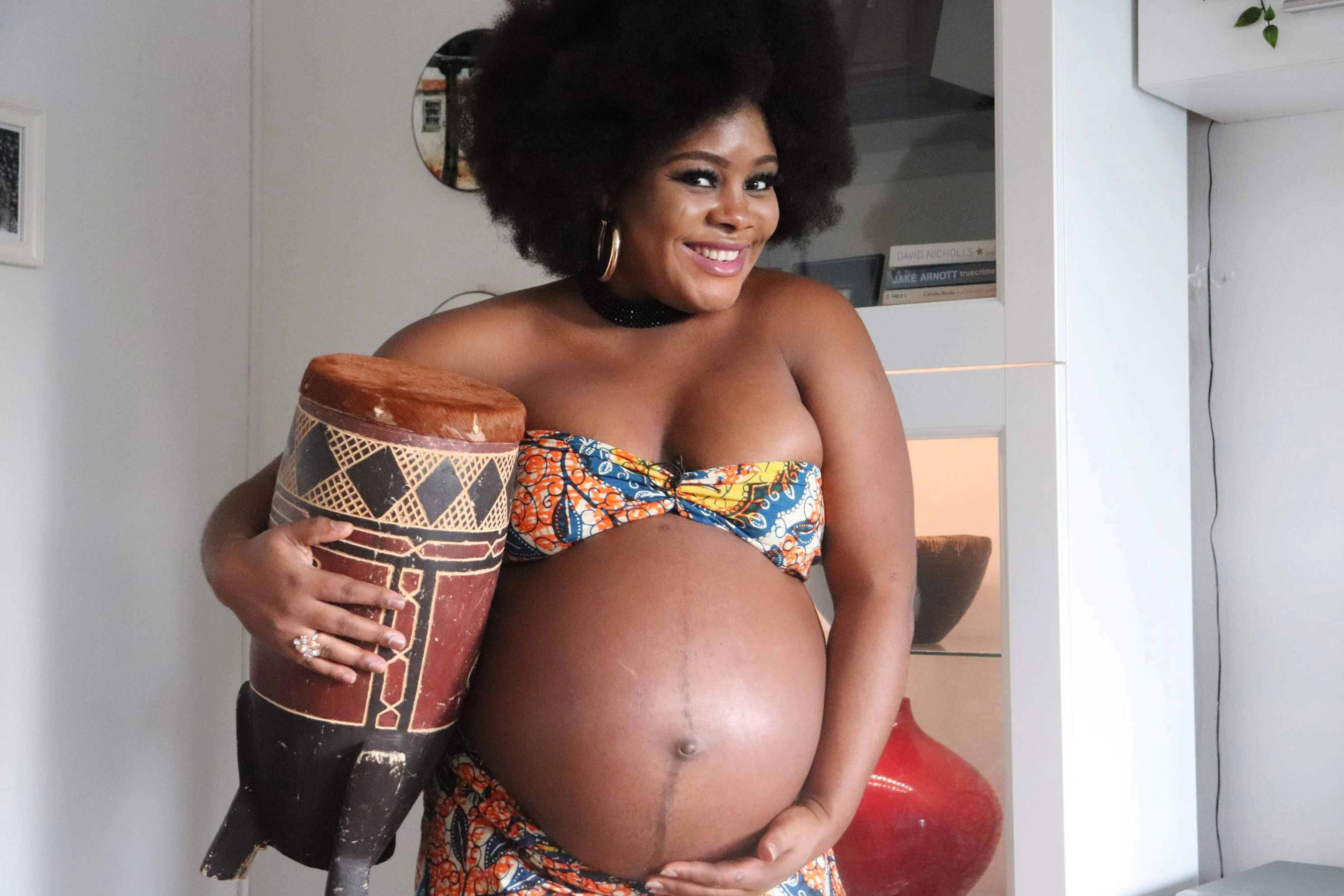 A pregnant woman in a patterned wrap standing in a warmly lit room, one hand resting on her side