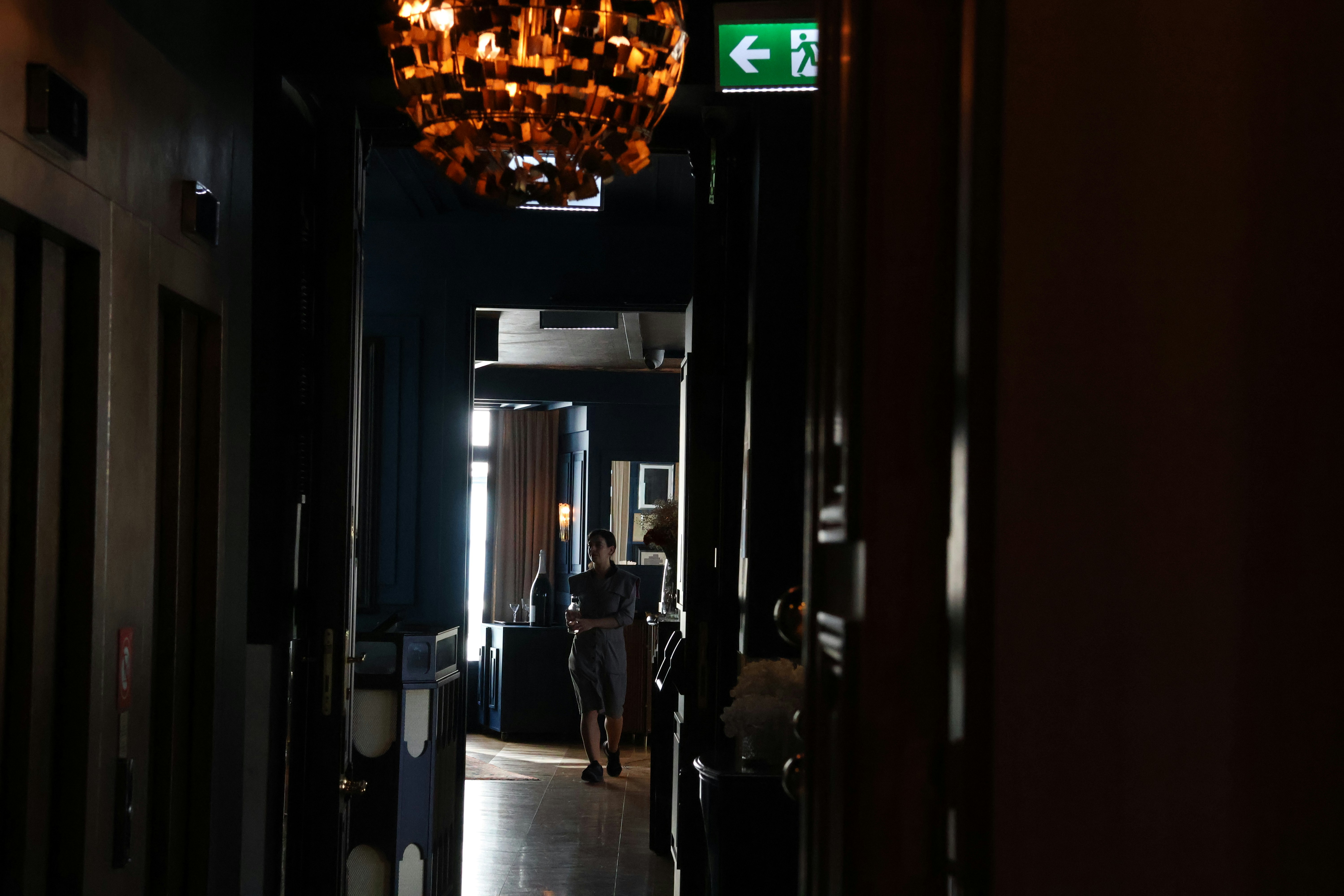 Dim, narrow hallway toward a bright doorway with a lone figure walking away. This photograph emphasizes depth, backlighting, and architectural contrast in a moody interior.