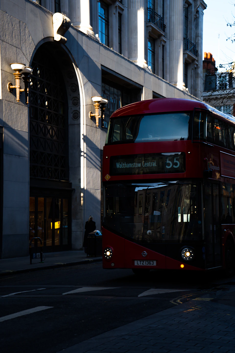 The image depicts a red double-decker bus parked on the side of a street, facing towards the right side of the frame. The bus is positioned next to an ornate white building with arched windows and a black awning, suggesting it may be located in London or another city with such architectural styles.

There are two people visible near the bus, possibly waiting for public transportation or walking along the sidewalk. A car can also be seen parked further down the street on the right side of the image.