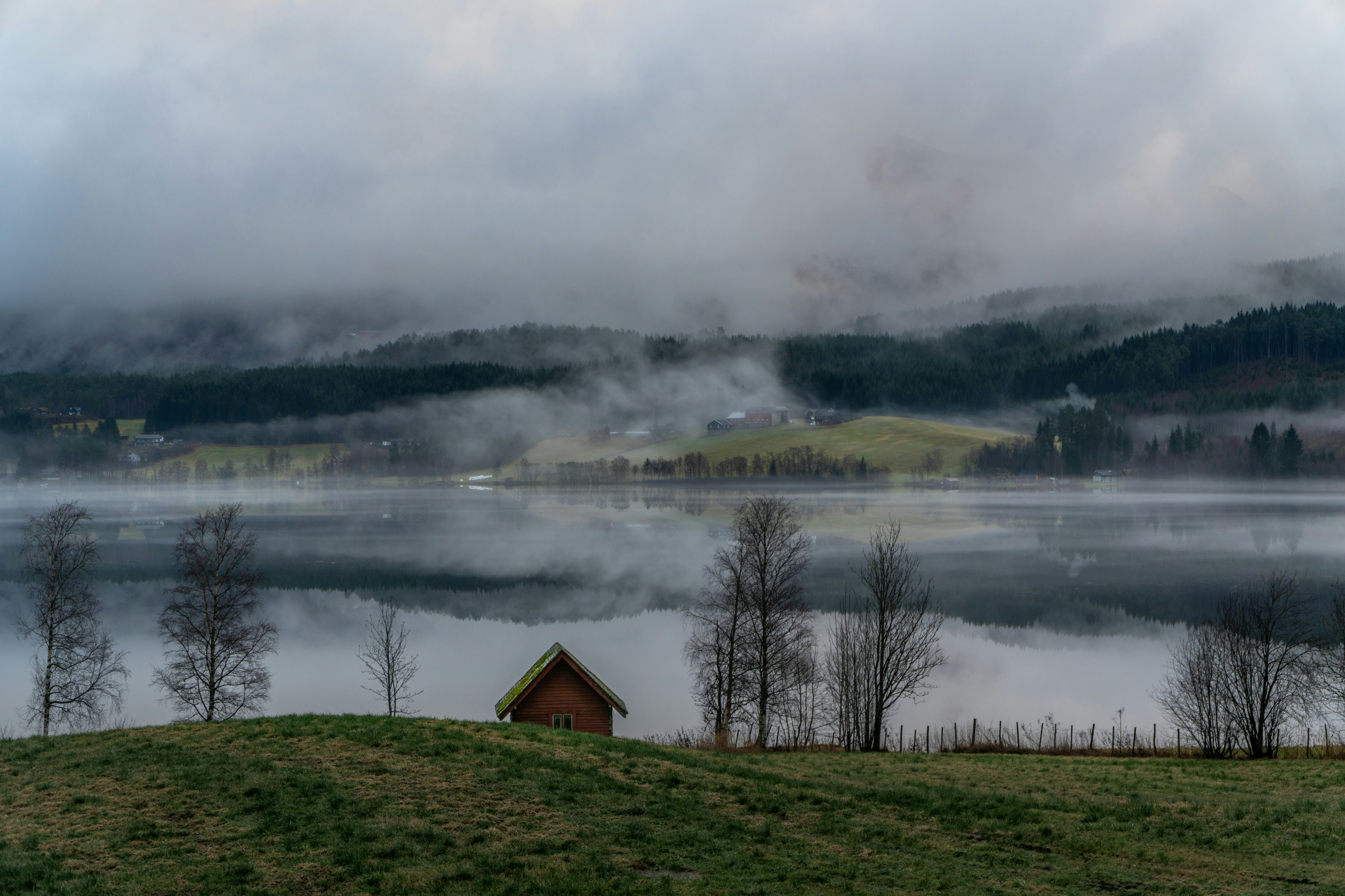 A small wooden hut sits in front of a misty lake and forestry mountains.