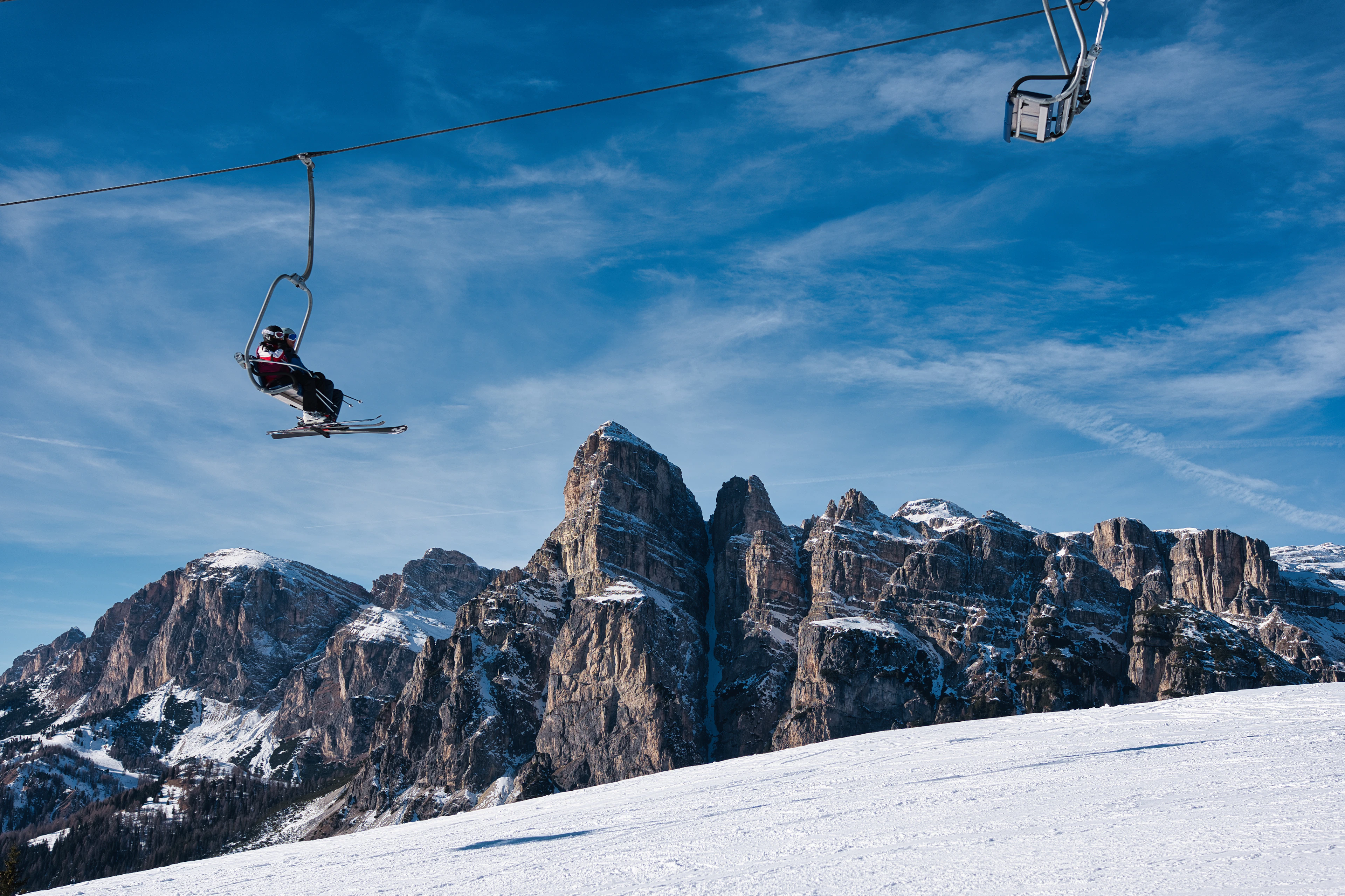 A skier rides a chairlift over a groomed slope beneath jagged rock peaks, skiing in Italy on a clear blue day