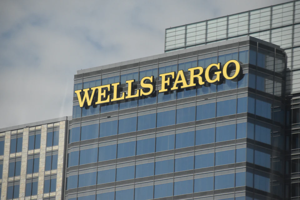 The image shows a large, modern glass and steel skyscraper with a prominent Wells Fargo sign on its side. The building is situated in an urban setting, surrounded by other structures that contribute to the cityscape. The perspective of the photo is from below, looking up at the towering height of the skyscraper against a cloudy sky.

The image does not contain any discernible text or additional objects, and there are no people visible in the frame.