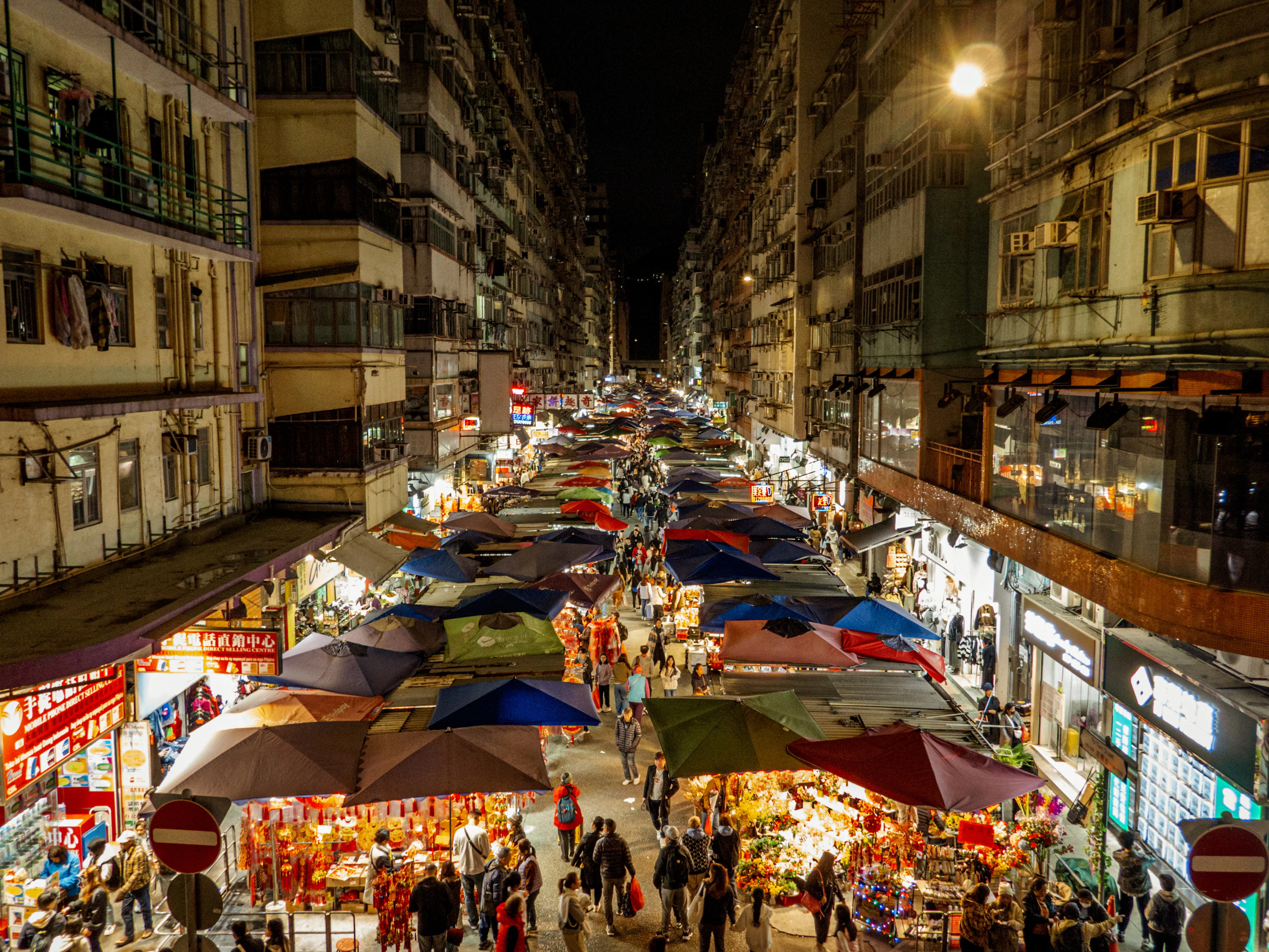 Crowds move between brightly coloured stall umbrellas on Fa Yuen Street market in Mong Kok after dark, lit shopfronts and tenement balconies rising on both sides of the lane.
