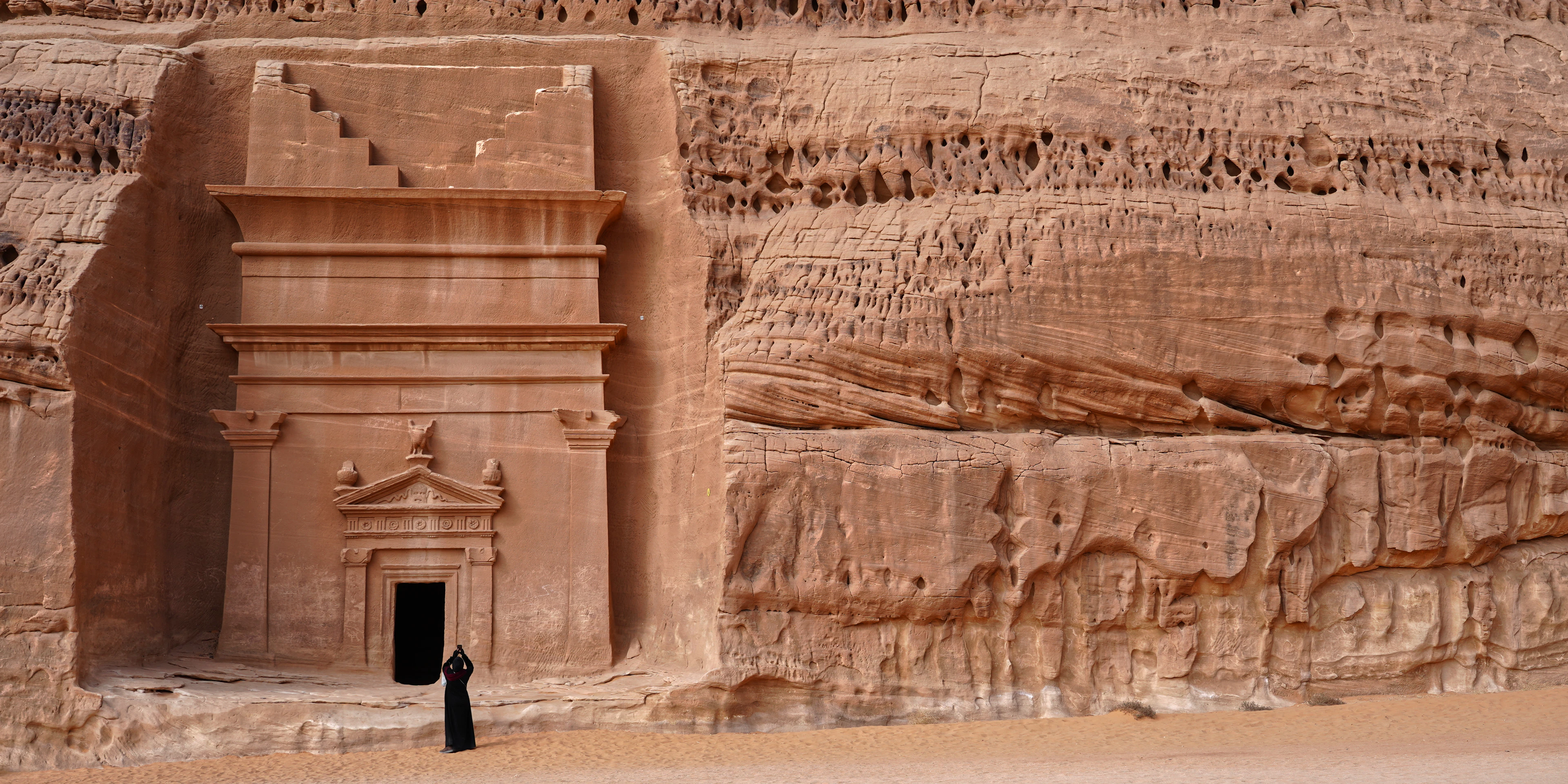 A lone visitor standing before a monumental Nabataean tomb facade carved into golden sandstone at Hegra, AlUla