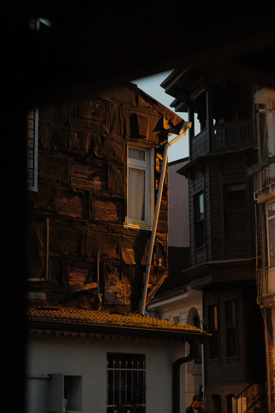 The image depicts a narrow street scene with two buildings on either side, one featuring a white window and the other a brown stone wall. The building on the left has a white window that reflects the warm glow of the setting sun, while the building on the right is partially obscured by its own window.

A person can be seen walking along the street in front of the buildings, adding a sense of life to the scene.