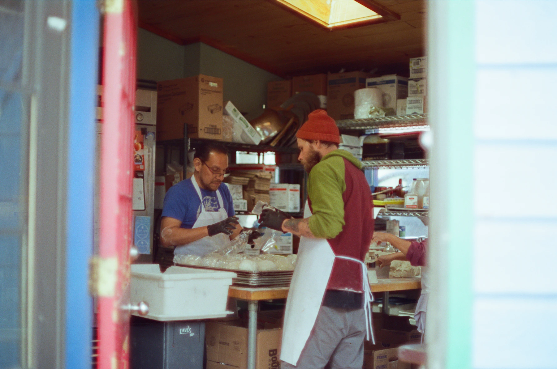 Dos personas con delantal preparando pedidos entre cajas, en la trastienda de un local