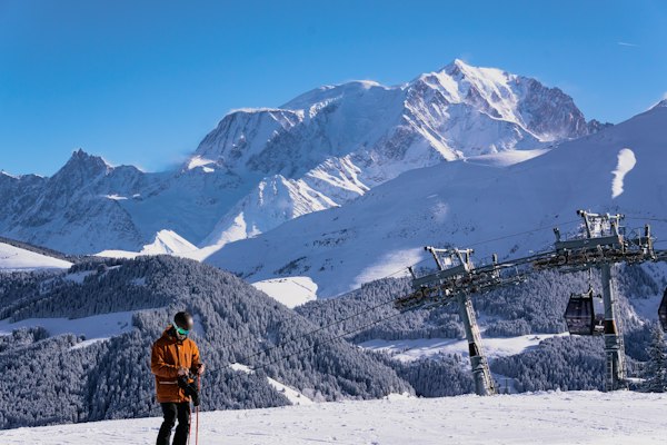 A skier standing at the top of a chairlift with the snow-covered Mont Blanc massif filling the skyline across the valley