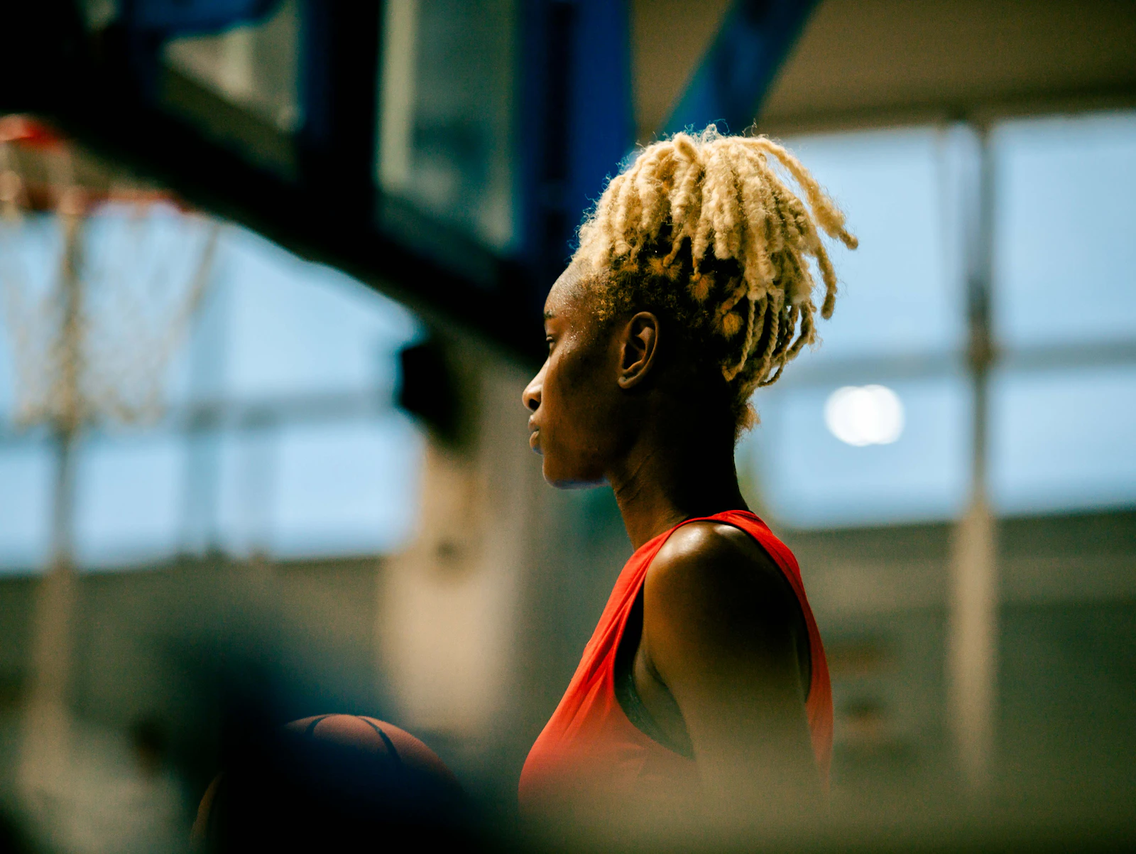 A woman with dreadlocks stands in quiet reflection in front of a basketball hoop.