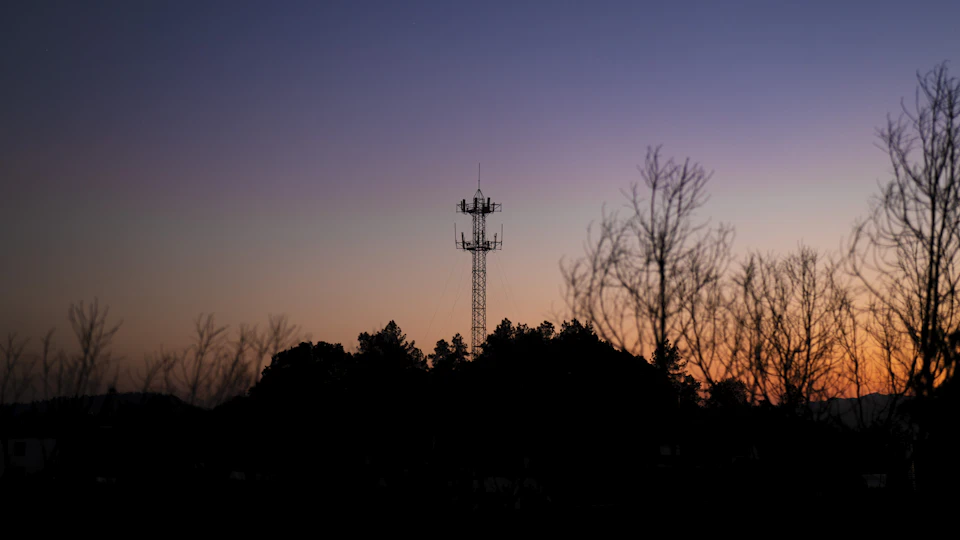 The image depicts a serene sunset over a rural landscape, with a tall tower standing out against the sky. The tower is surrounded by trees and bushes, creating a sense of tranquility and solitude. The colors in the sky are predominantly purple and orange, reflecting off the warm hues of the setting sun. The low angle of the shot enhances the tower's prominence within the scene, making it appear as if it reaches towards the heavens.