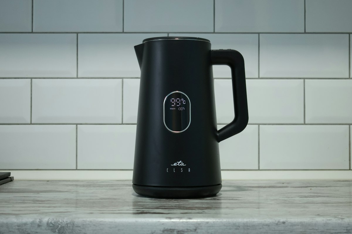 Pava eléctrica negra mate con visor digital que marca 99 °C, apoyada sobre su base en una mesada de mármol gris frente a una pared de azulejos blancos tipo subte