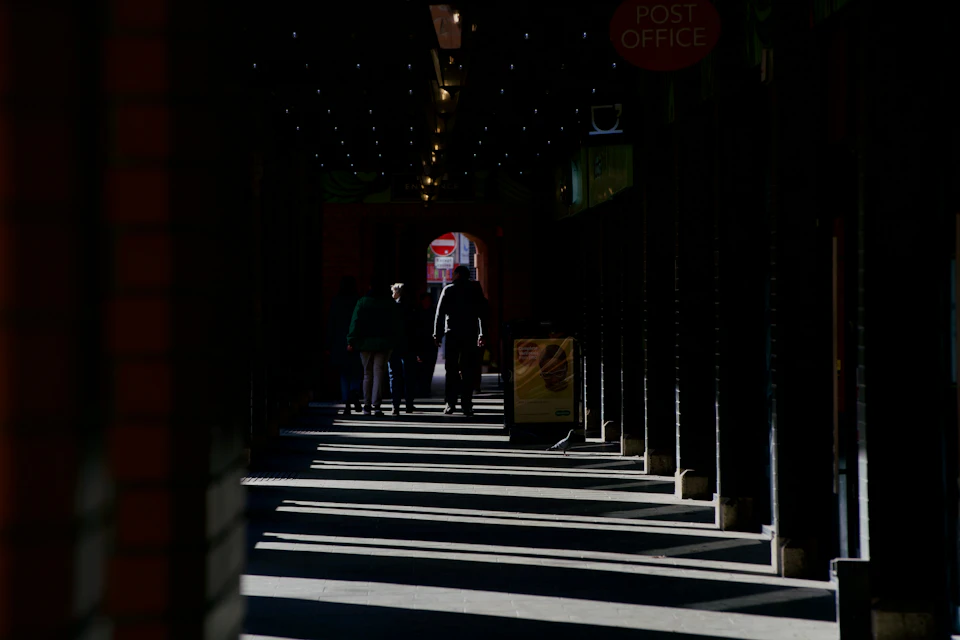 The image depicts a group of people walking down a long, narrow hallway with tall columns on either side. The hallway is illuminated by natural light streaming through windows on both sides, creating a warm and inviting atmosphere. On one side of the hallway, there are two individuals wearing dark clothing, while on the other side, three more people can be seen in lighter attire. The scene suggests that they might be walking towards an office building or another type of establishment with columns lining its exterior.