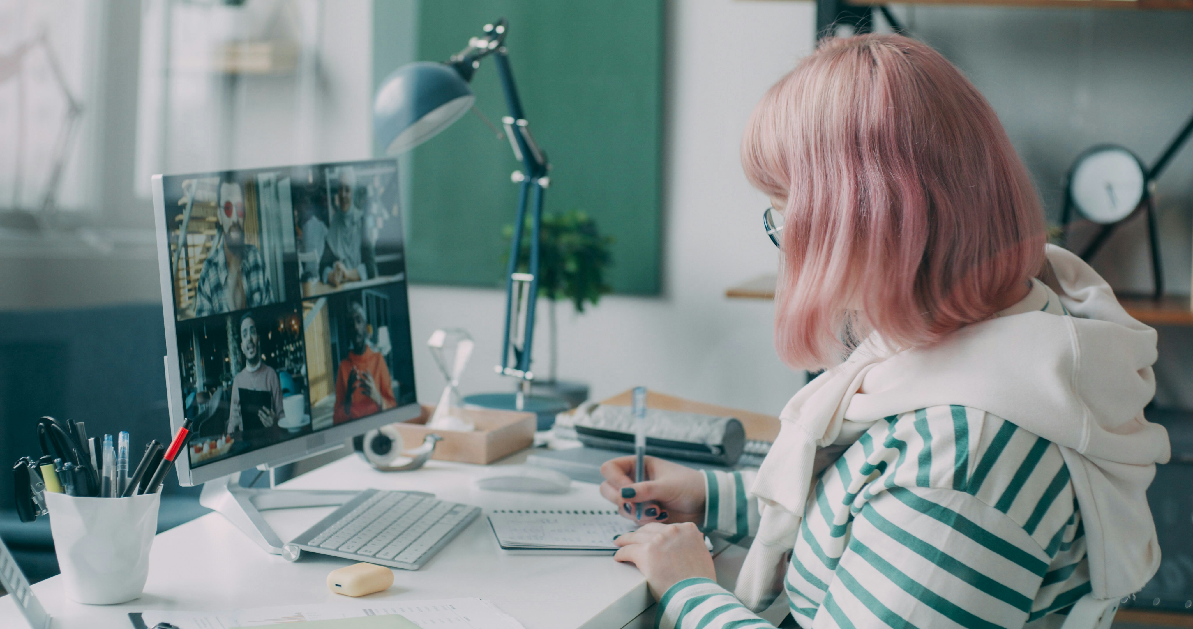 Person with pink hair takes notes during a video conference.