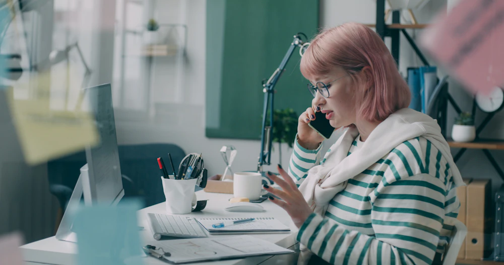 A woman at an office desk on the phone, with notes and a computer in front of her