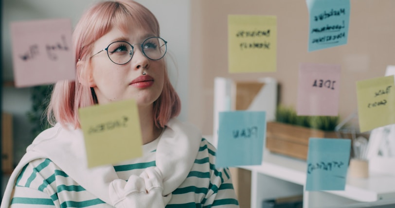 A woman with pink hair and glasses standing in front of post it notes