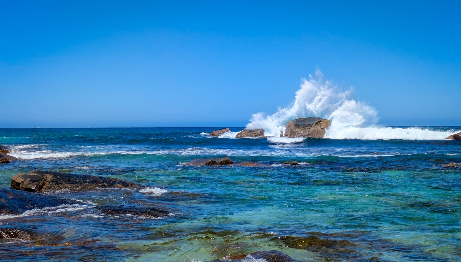 A picture of the ocean at a beach. In the foreground are some vibrant shallows interspersed with rocks. The water is so clear and colourful it is almost green in places. Just right of the centre is a large rock being smashed from behind by water, sending a wave up into the air. In the background is a very clear blue sky.