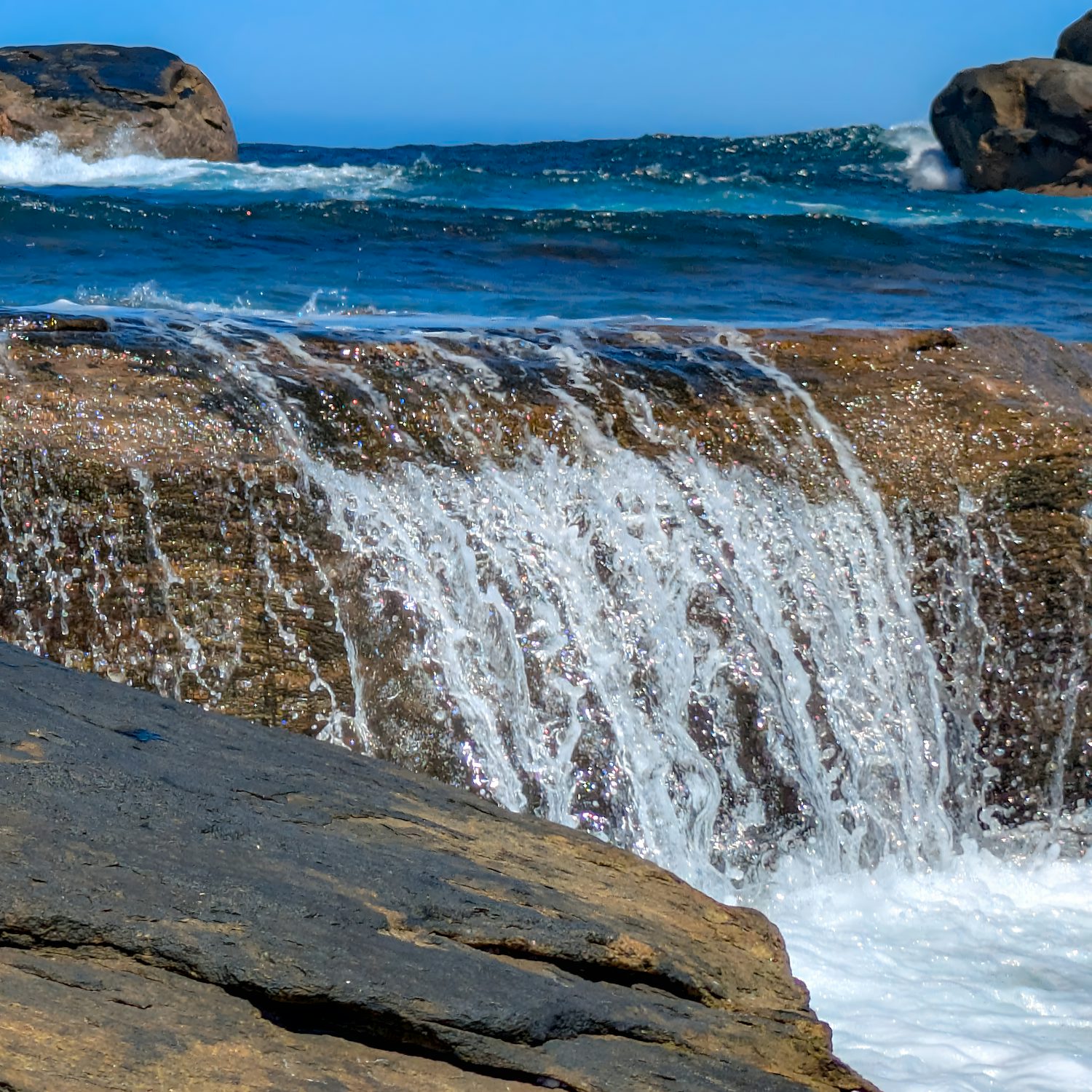 Roiling water cascading over the side of a rock. Behind it is the turbulent blue sea, with the clear, lighter blue sky behind it.