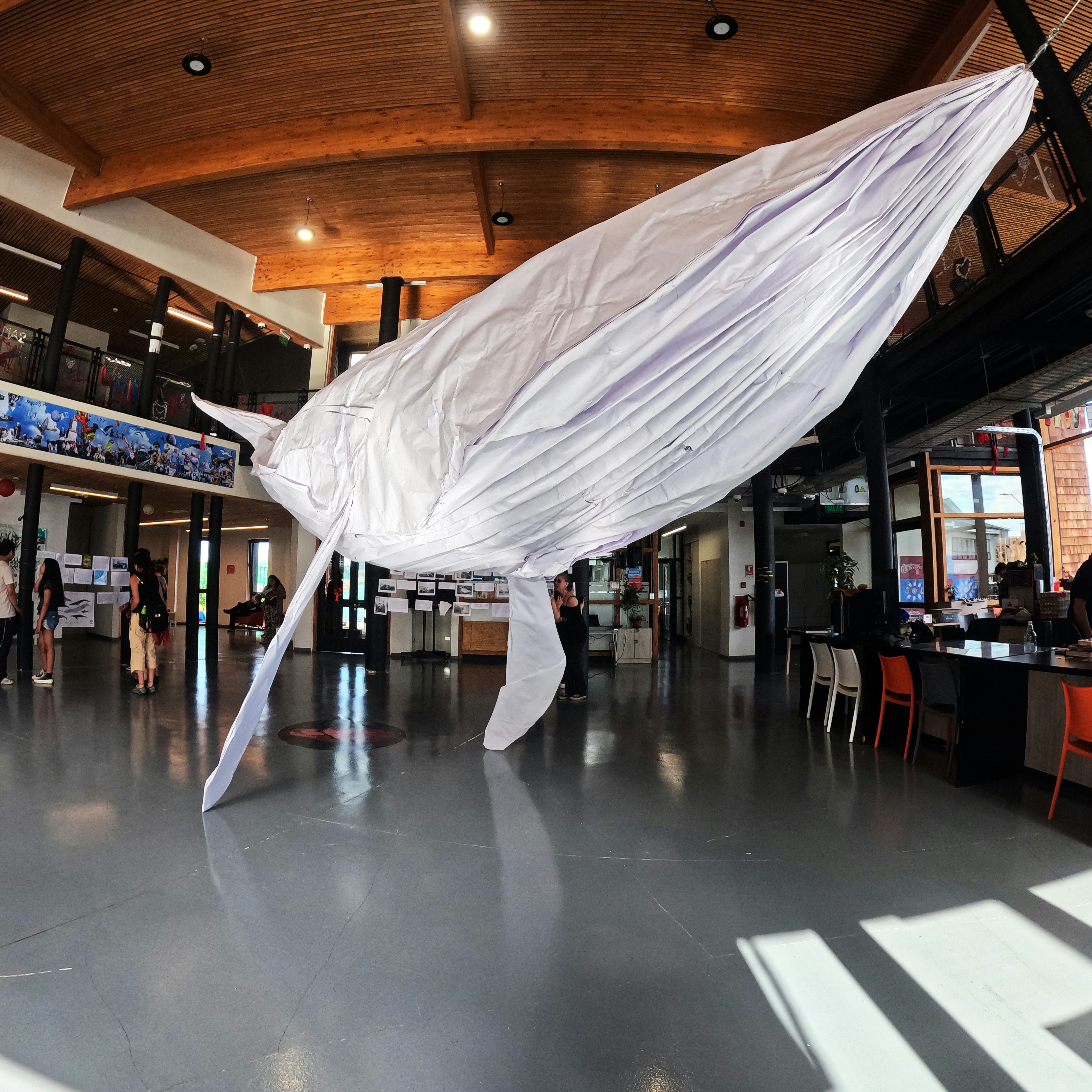 A large white fabric sheet billows mid-air inside a spacious interior with wooden ceilings and a polished concrete floor, suggesting a performance or installation in a public venue.